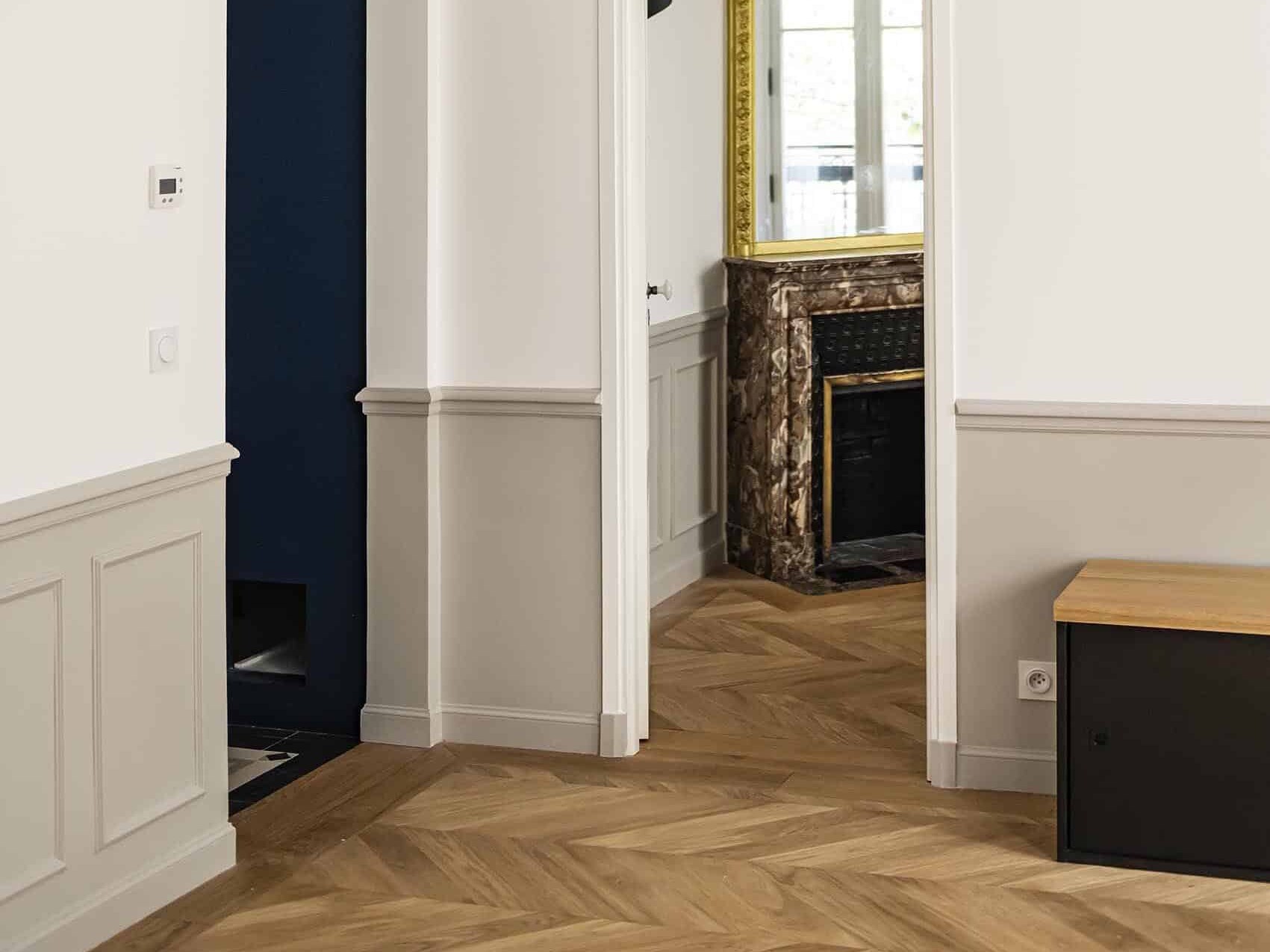 Vue intérieure montrant un couloir avec un parquet en bois à chevrons, des murs blancs et un aperçu d'une pièce avec une cheminée et une fenêtre.