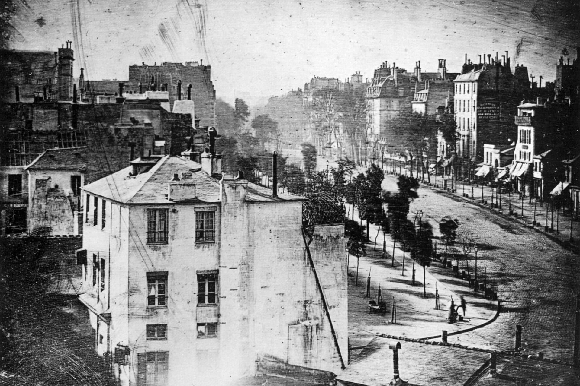 Photographie en noir et blanc d'une rue de la ville avec plusieurs immeubles à plusieurs étages, des arbres bordant le trottoir et quelques personnes visibles dans la rue autrement vide.
