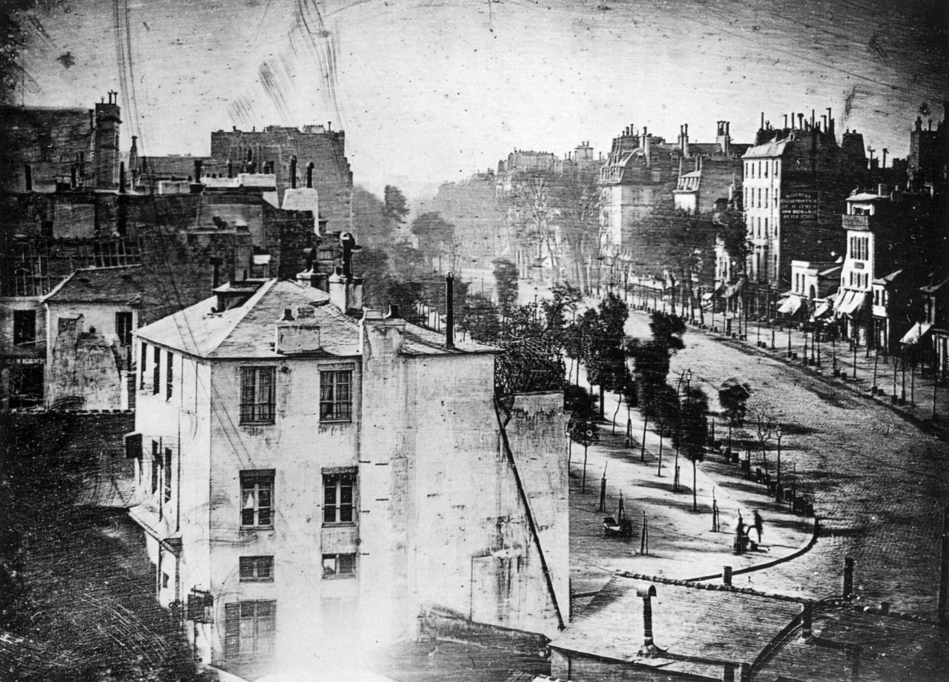 Photographie en noir et blanc d'une rue de la ville avec plusieurs immeubles à plusieurs étages, des arbres bordant le trottoir et quelques personnes visibles dans la rue autrement vide.
