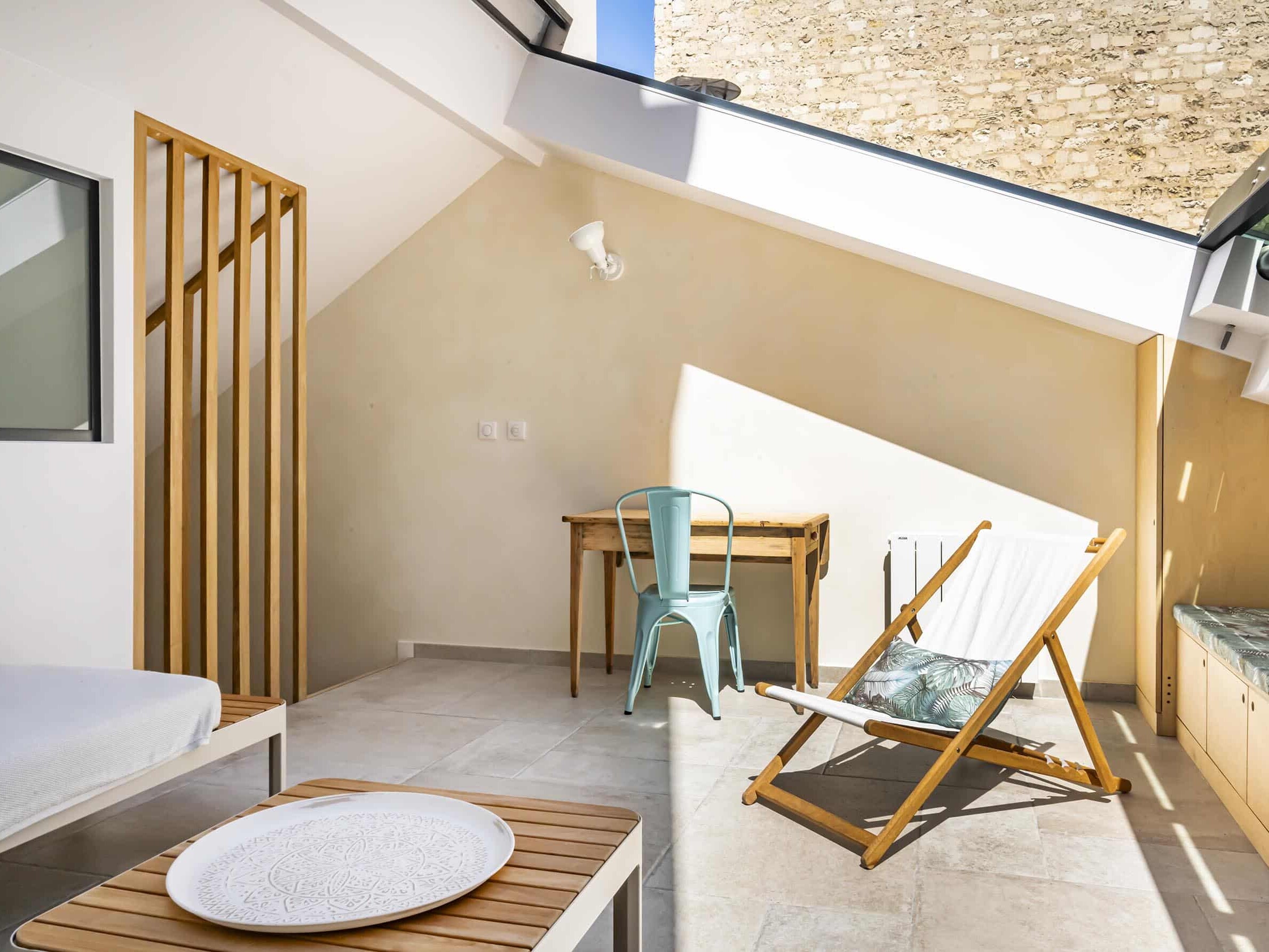 Une pièce ensoleillée avec un bureau, une chaise en métal vert, une chaise longue, un banc, une petite table avec une assiette décorative et un sol carrelé beige.