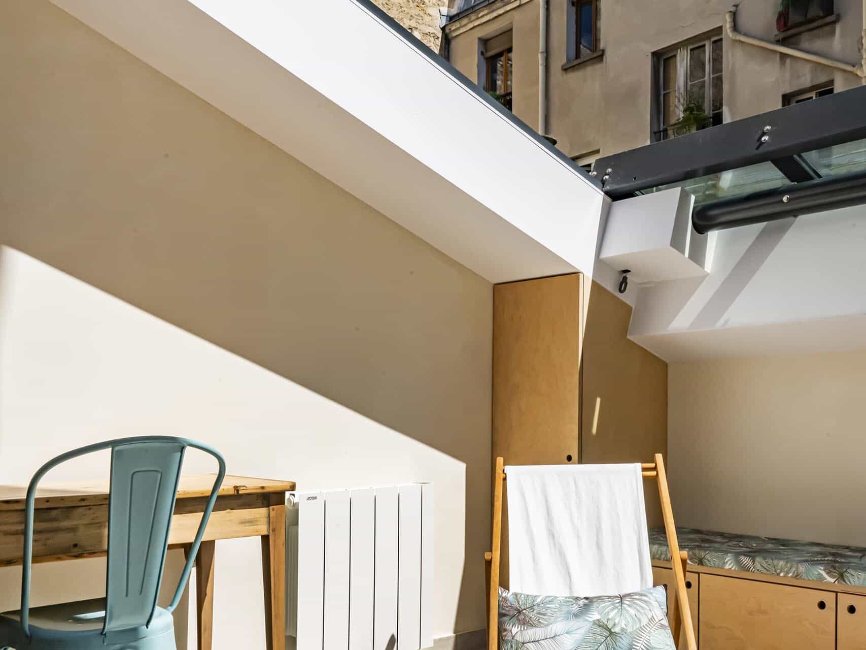 Une petite terrasse sur le toit avec un bureau en bois et une chaise en métal, une chaise longue pliante avec un coussin, des murs beiges et une vue sur les bâtiments voisins.