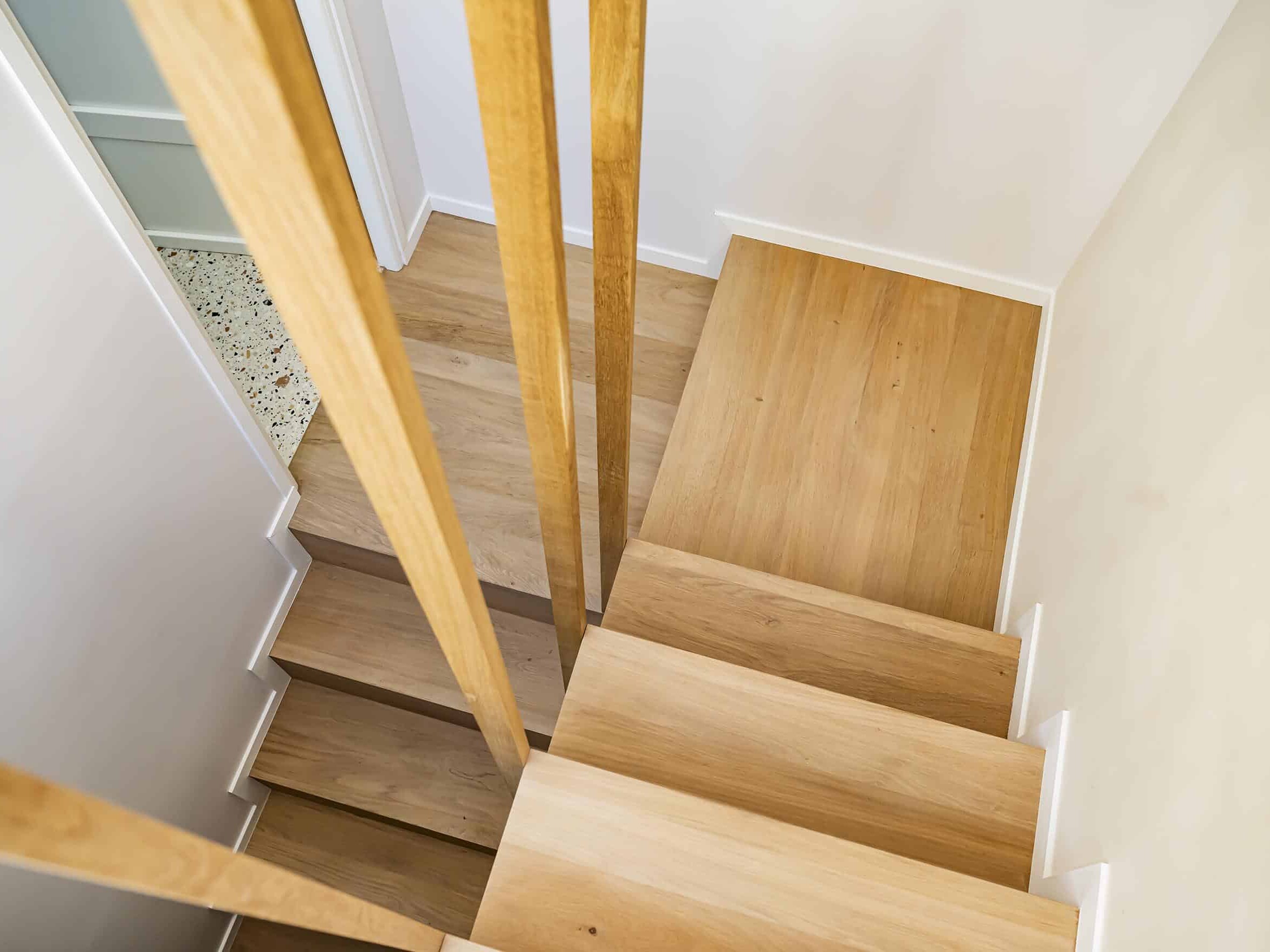 Escalier en bois avec rampes verticales en bois vu de dessus, comportant des marches en bois de couleur claire et un palier, avec des murs blancs des deux côtés.