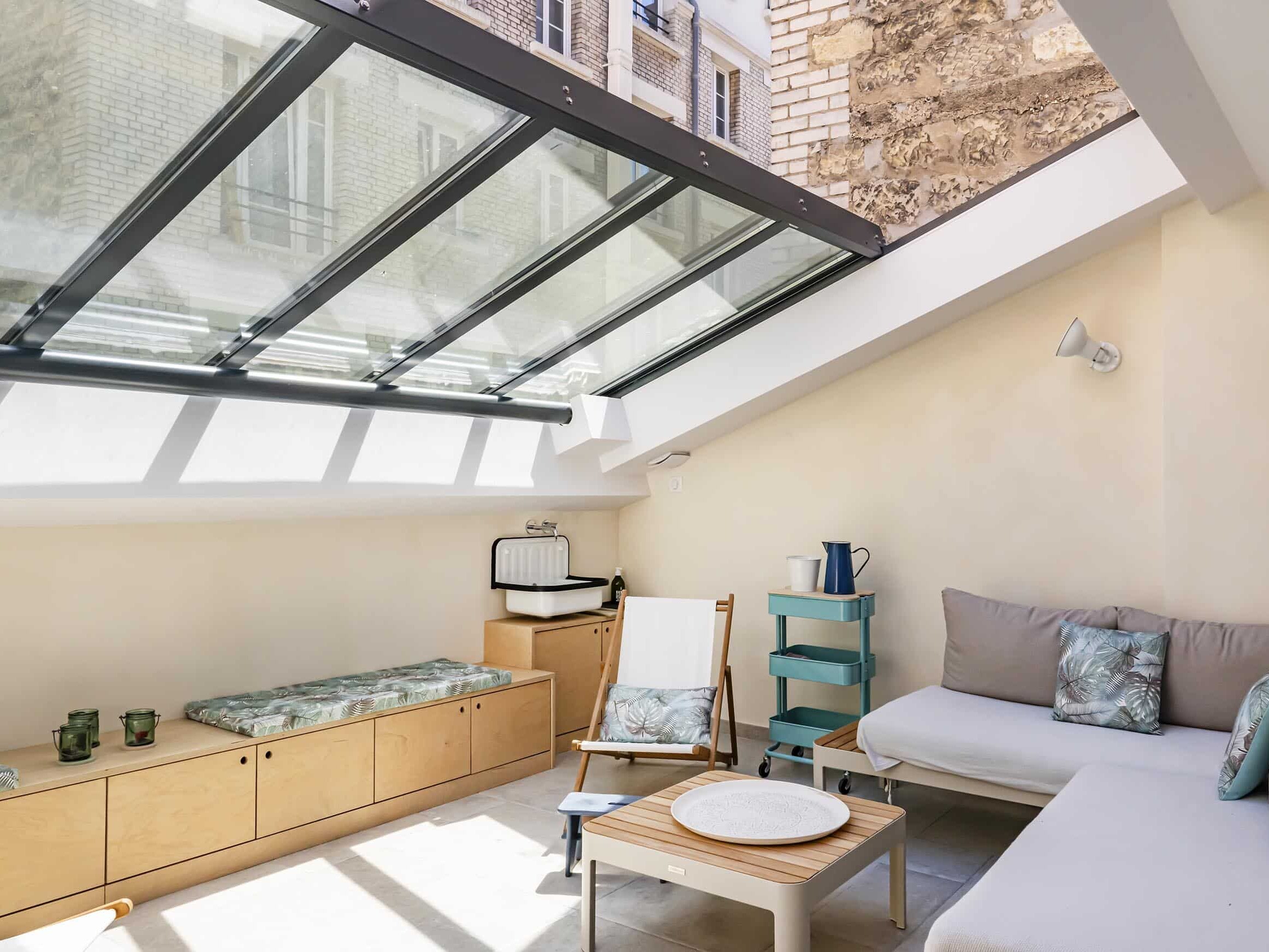 Un salon intérieur ensoleillé avec un toit en verre, des meubles en bois clair, un canapé gris, une chaise longue, des bancs de rangement et une petite table basse.