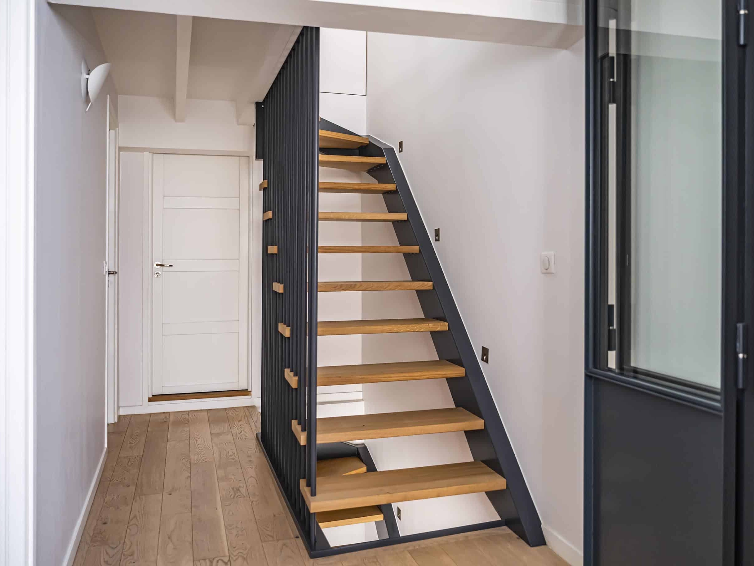 Un couloir moderne avec parquet, un escalier ouvert en métal noir et bois, des murs blancs et une porte blanche fermée au bout.