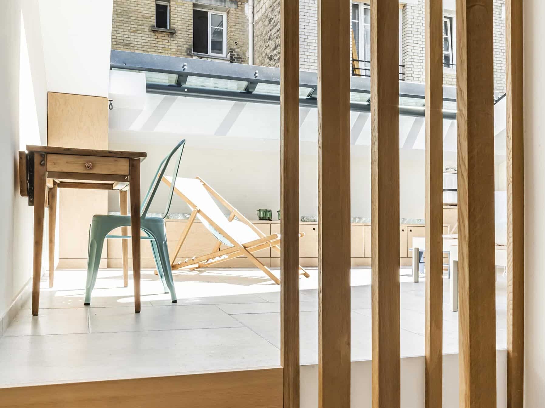 Des escaliers en bois mènent à un petit espace de travail avec un bureau et une chaise en métal, à côté d'une terrasse extérieure lumineuse avec une chaise longue ; les fenêtres de l'immeuble sont visibles à l'extérieur.