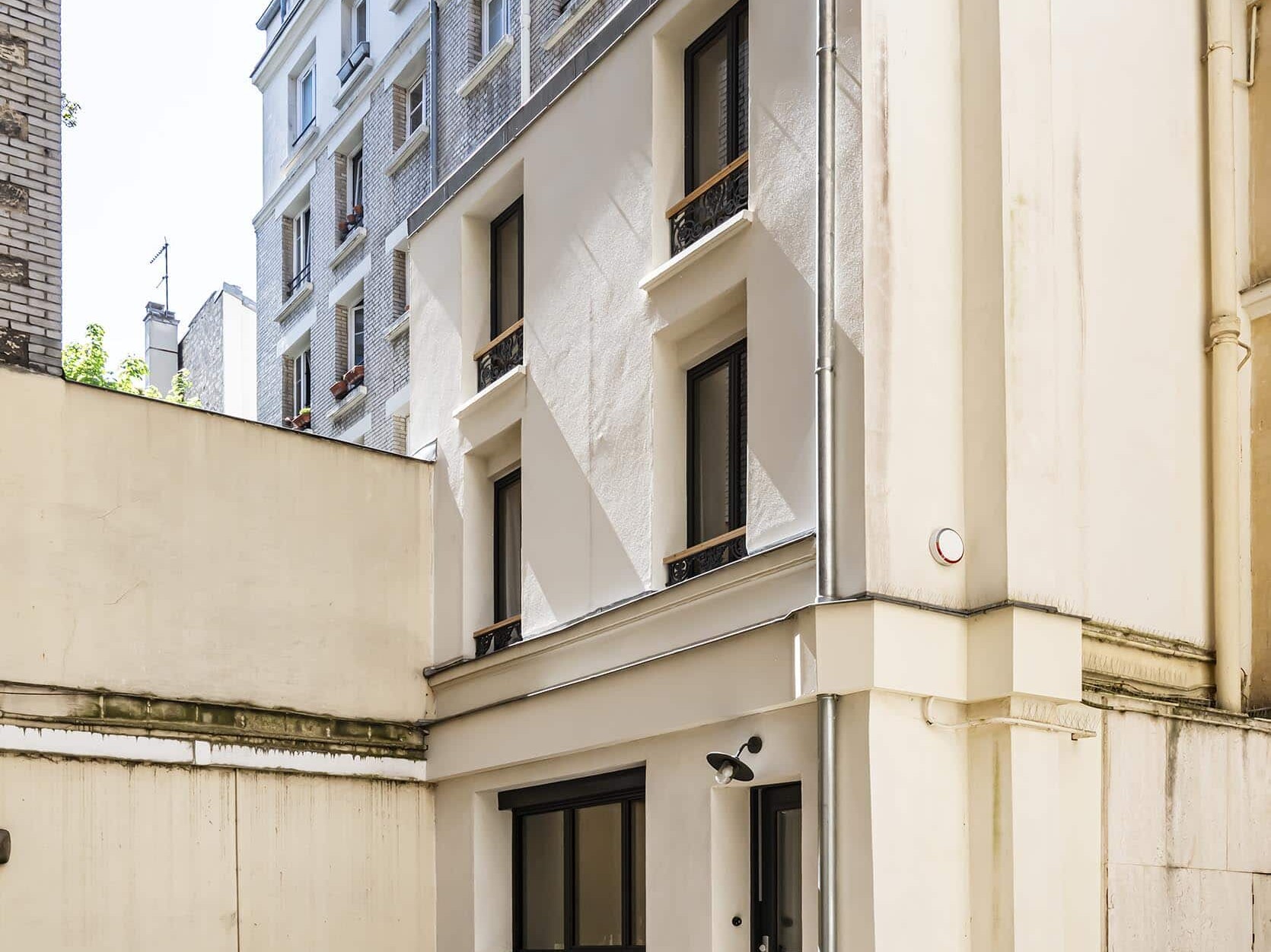 Une petite maison étroite de trois étages avec une façade beige se dresse dans une cour pavée à Paris, entourée de bâtiments plus hauts ; une moto couverte et des plantes en pot se trouvent près de l'entrée.