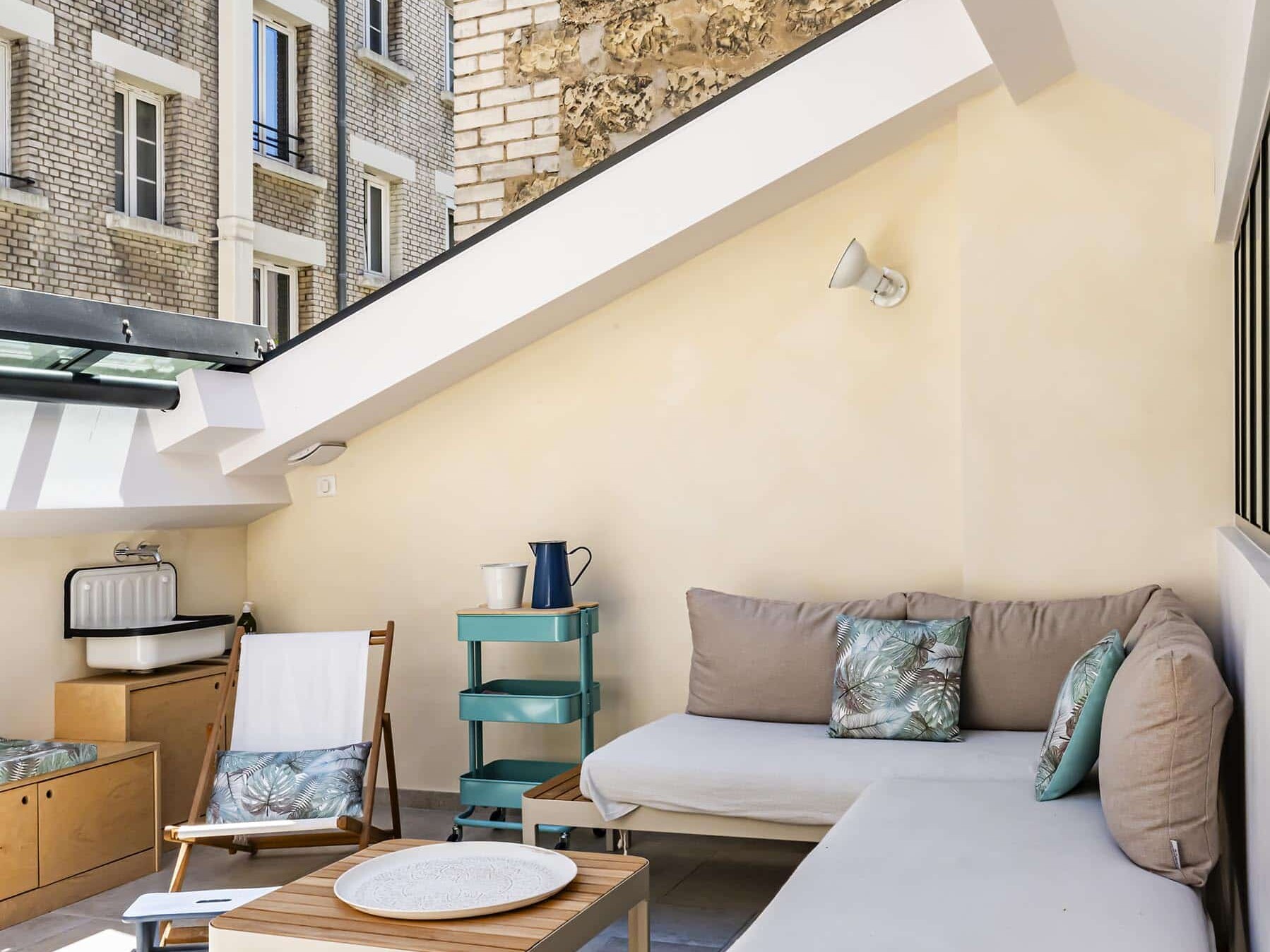 Un balcon moderne avec un banc rembourré en forme de L, une table basse en bois, une chaise longue et des coussins décoratifs sous un toit en pente, donnant sur les bâtiments voisins.