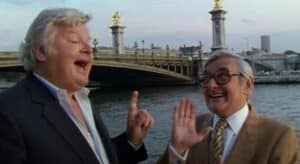 Deux hommes âgés se tiennent debout et font des gestes animés devant un pont sur une rivière, avec des lampadaires ornés visibles en arrière-plan.