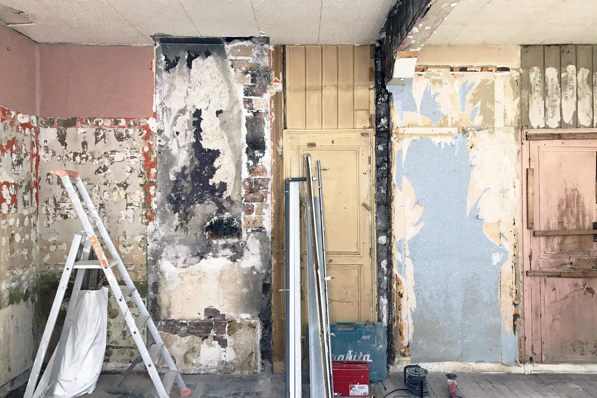Chambre en rénovation avec murs apparents et abîmés, outils de construction, une échelle et des poutres métalliques appuyées contre le mur.