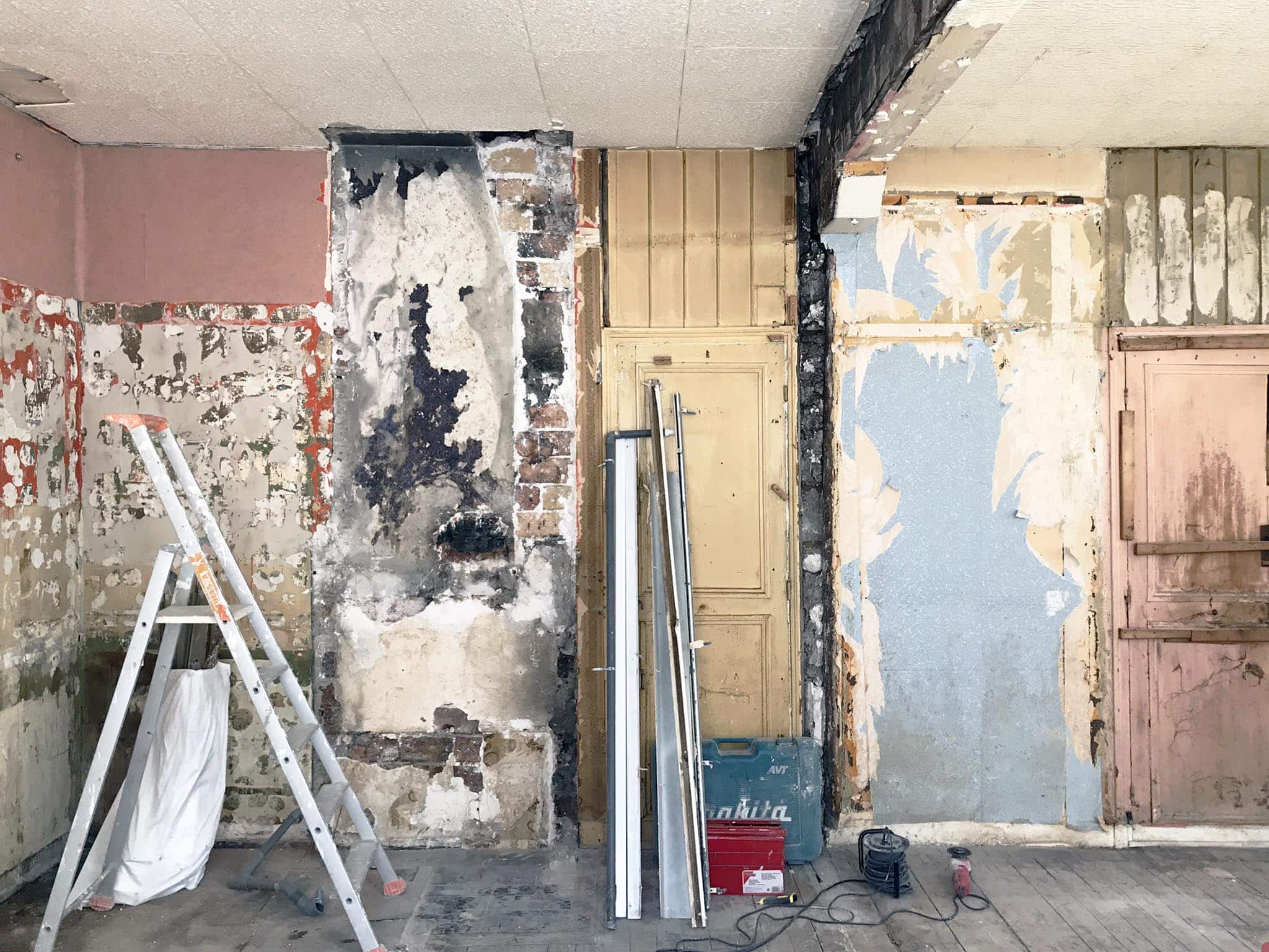 Chambre en rénovation avec murs apparents et abîmés, outils de construction, une échelle et des poutres métalliques appuyées contre le mur.