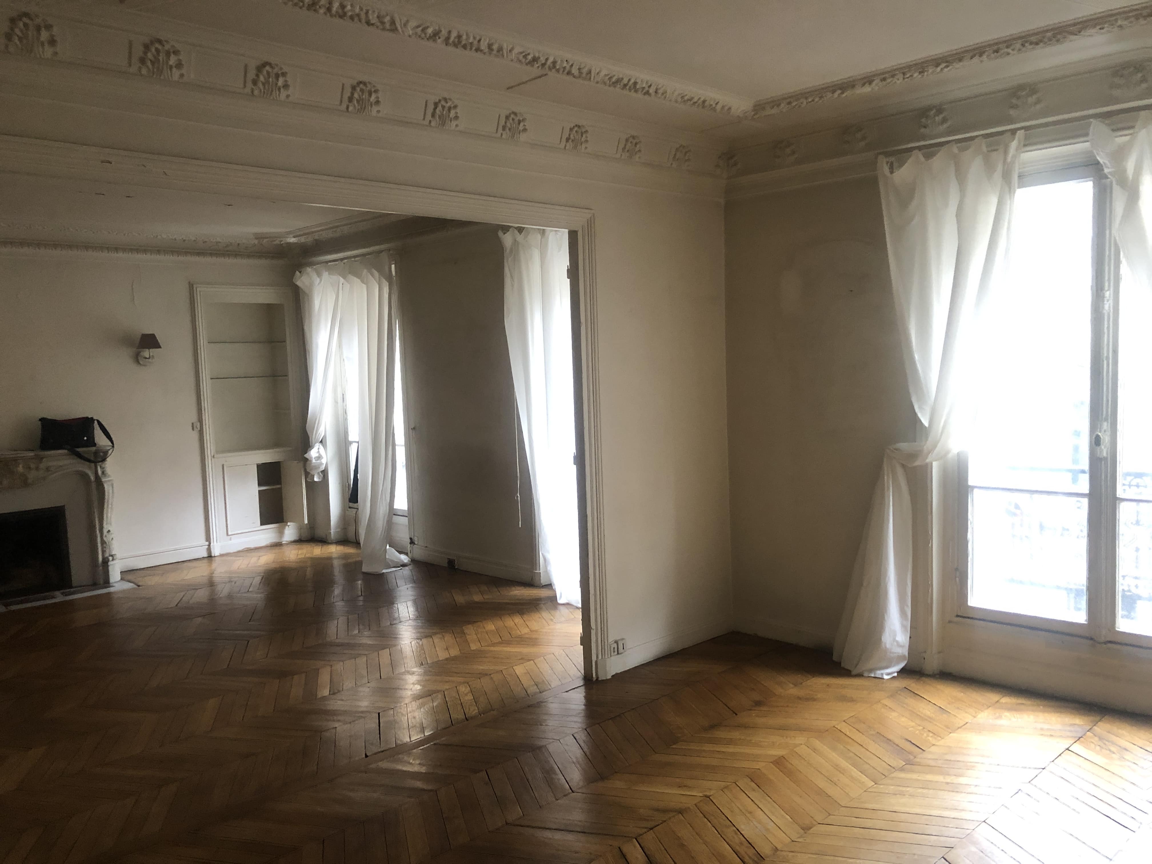 Pièce vide avec un sol en bois à chevrons, des murs blancs, de grandes fenêtres avec des rideaux transparents, des moulures décoratives au plafond et une cheminée sur la gauche.