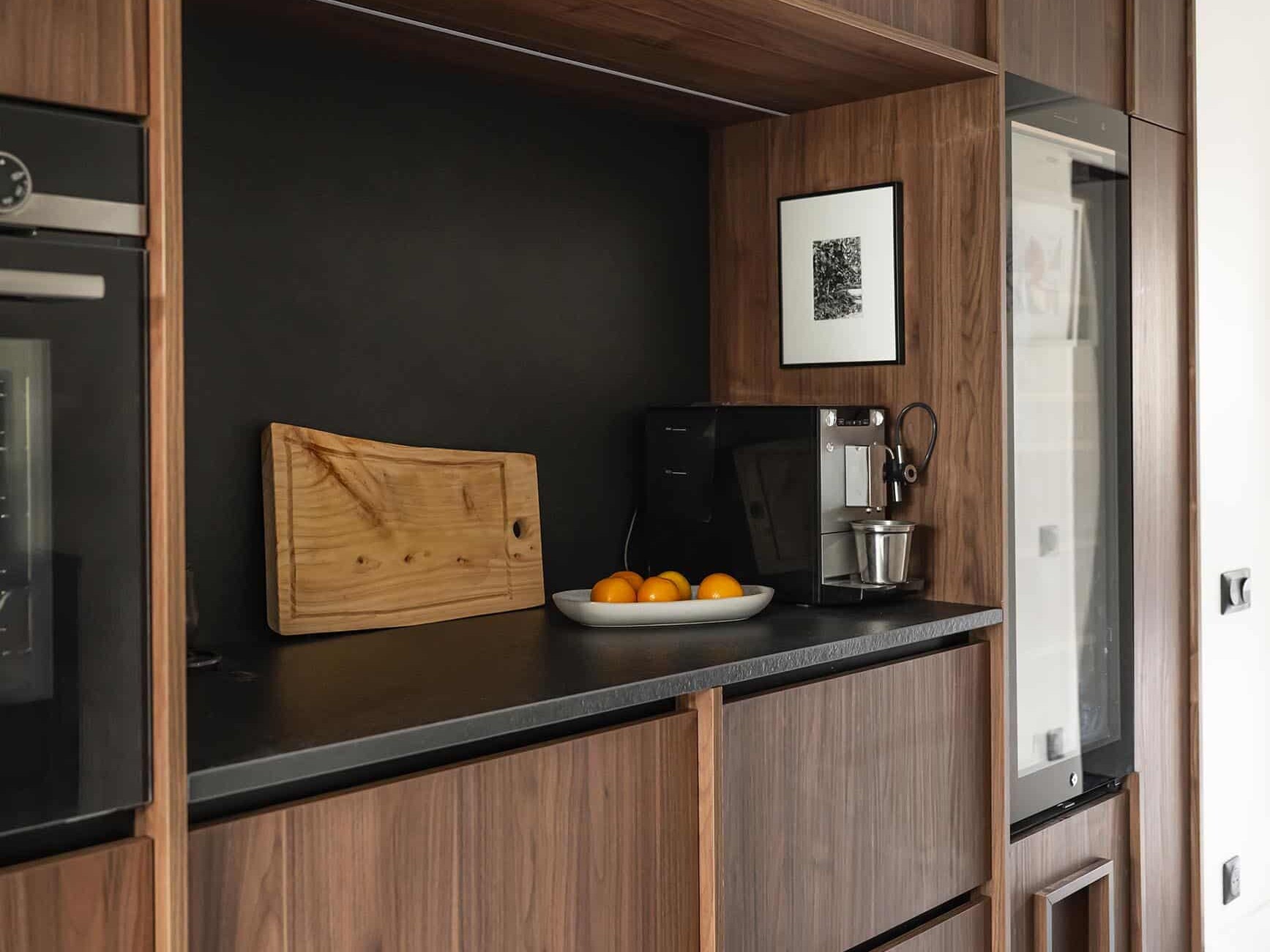 Cuisine moderne avec meubles en bois foncé, comptoir noir, planche à découper, bol d'oranges, cafetière, mixeur et œuvre d'art encadrée sur l'étagère du dosseret.