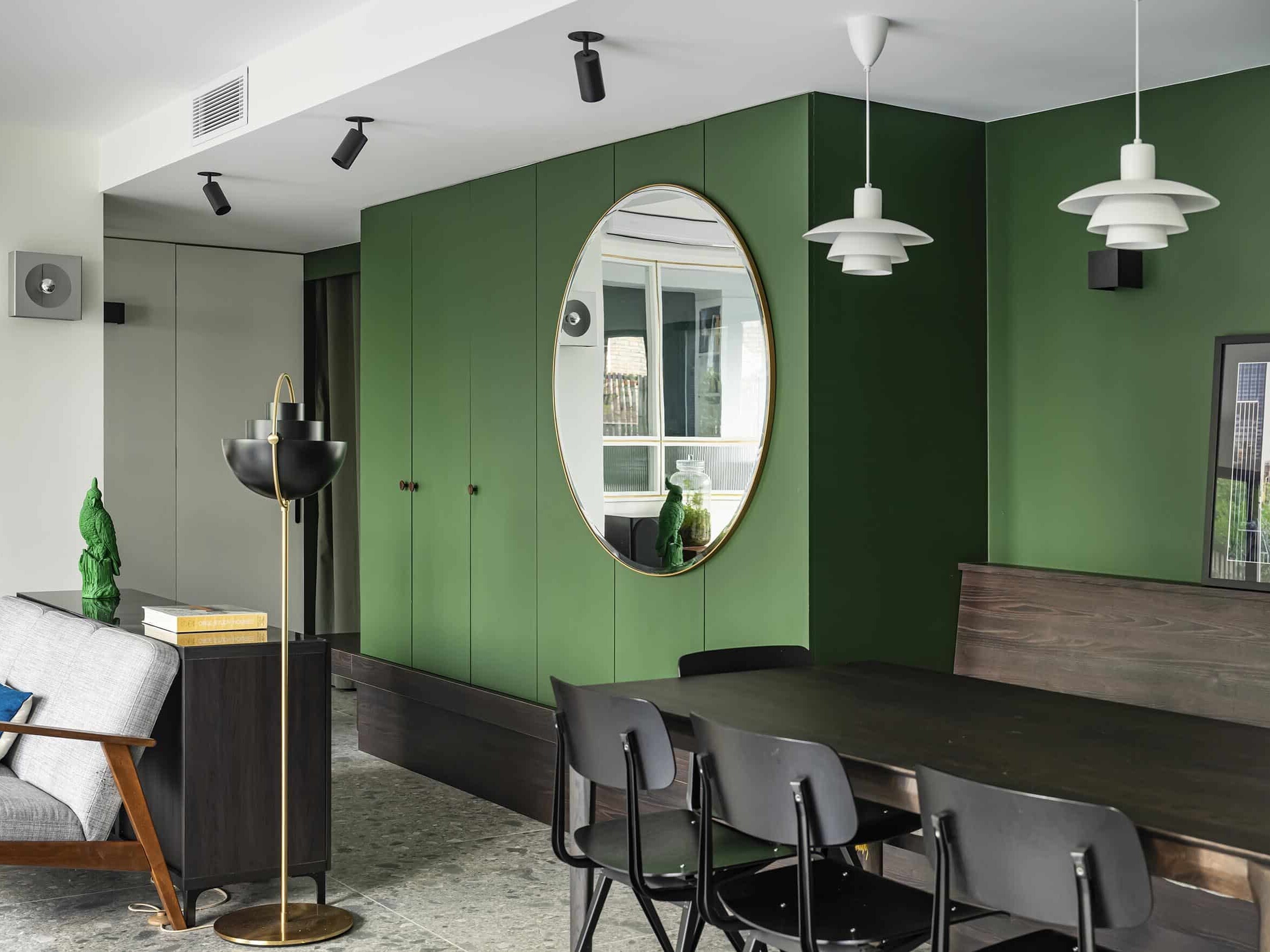 Salle à manger moderne avec armoires vertes, miroir mural rond, table et chaises de salle à manger noires, lampes suspendues et fauteuil gris avec table d'appoint.