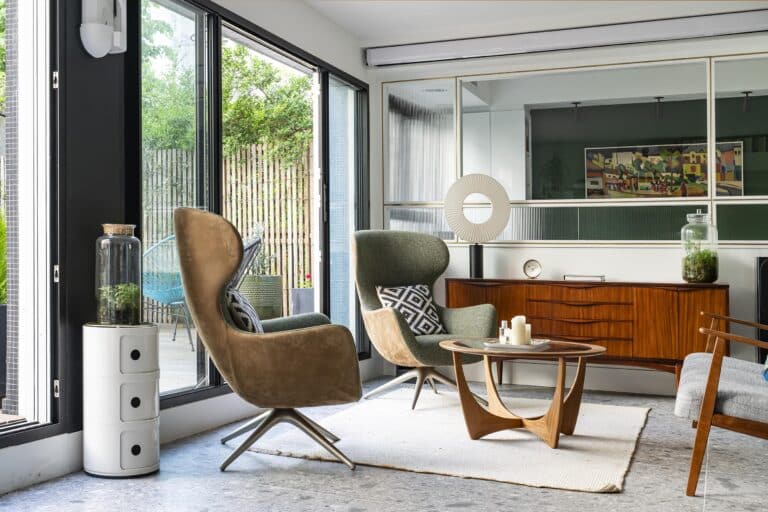Un salon moderne avec de grandes fenêtres, deux fauteuils, une table basse en bois, un buffet, des objets de décoration et des plantes, avec un patio visible à l'extérieur.