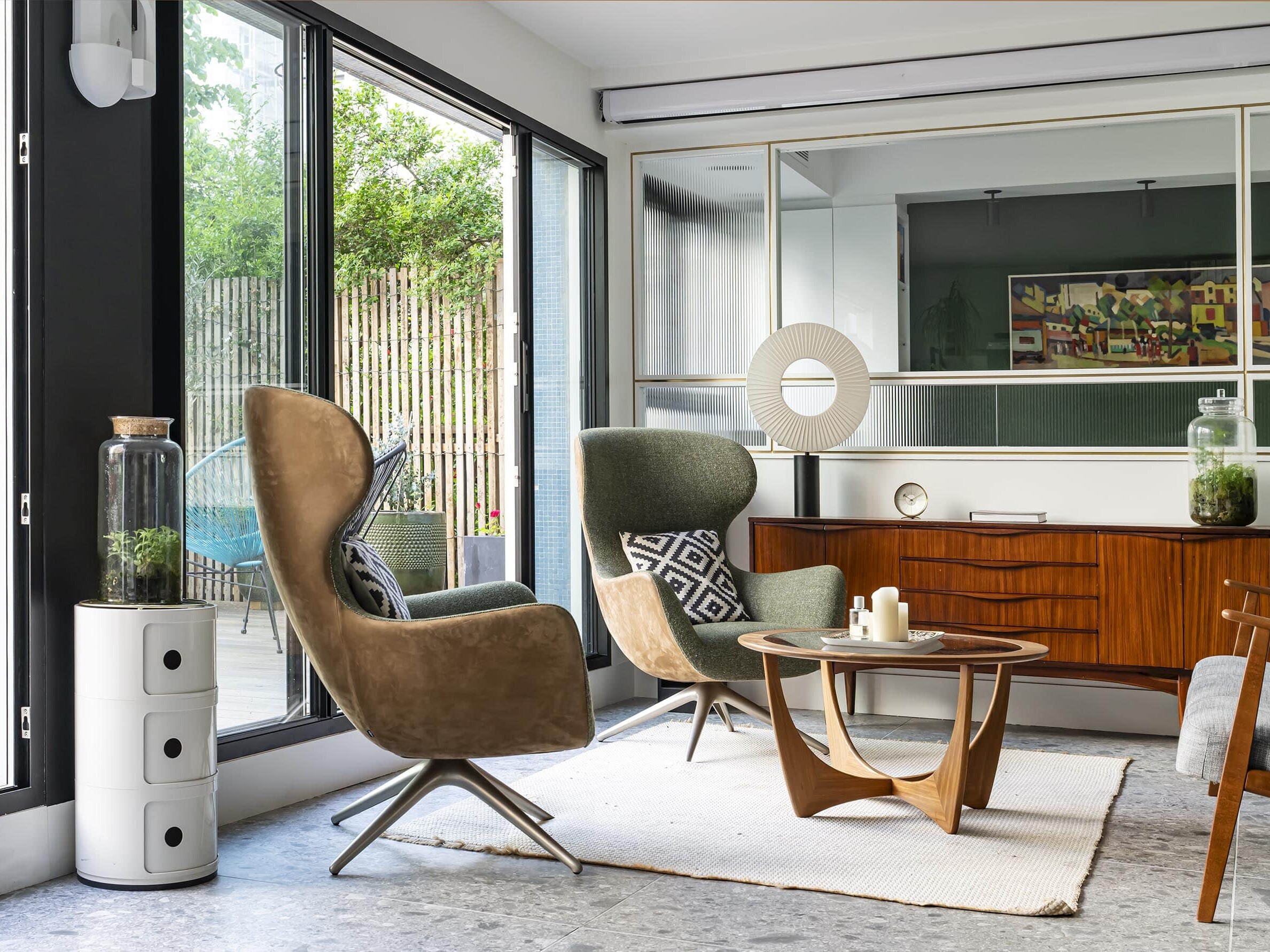 Un salon moderne avec de grandes fenêtres, deux fauteuils, une table basse en bois, un buffet, des objets de décoration et des plantes, avec un patio visible à l'extérieur.