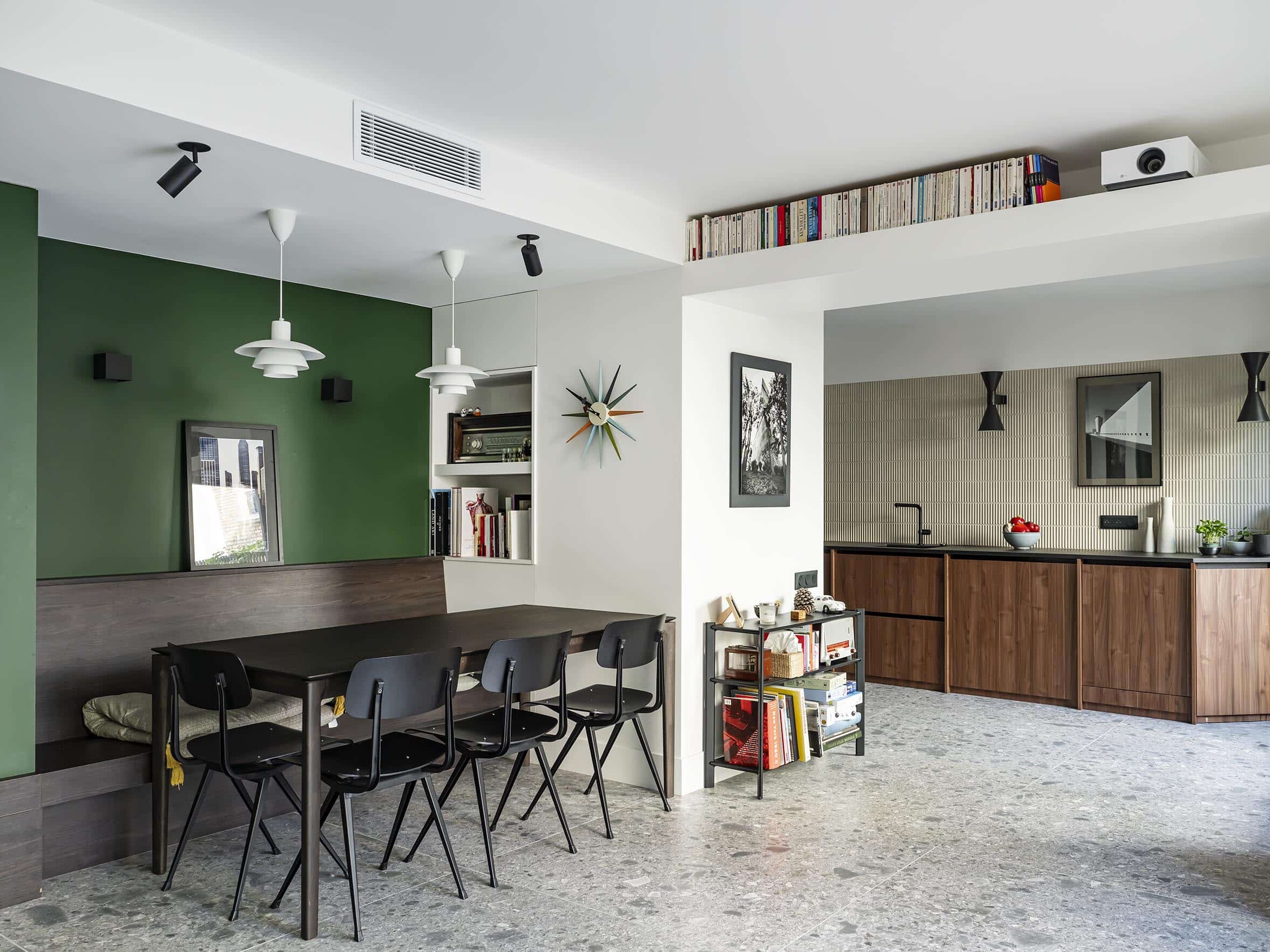 Salle à manger et cuisine moderne ouverte avec un mur d'accent vert, des chaises noires autour d'une table foncée, des armoires en bois, des étagères murales et une bibliothèque au-dessus de la cuisine.