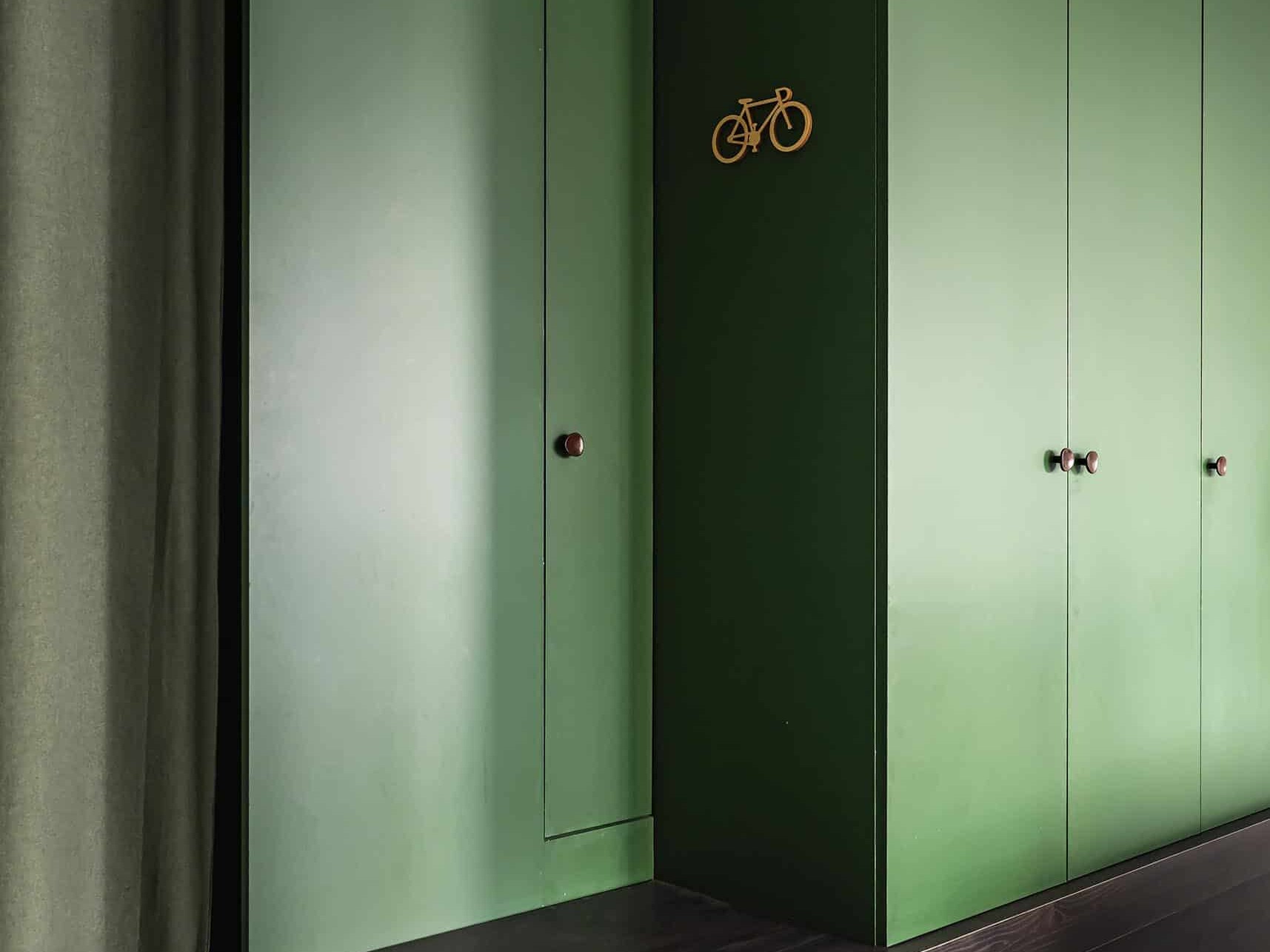 Meubles encastrés verts avec banquette en bois foncé et petite bicyclette dorée au mur ; sol en carreaux de pierre et rideau à gauche.