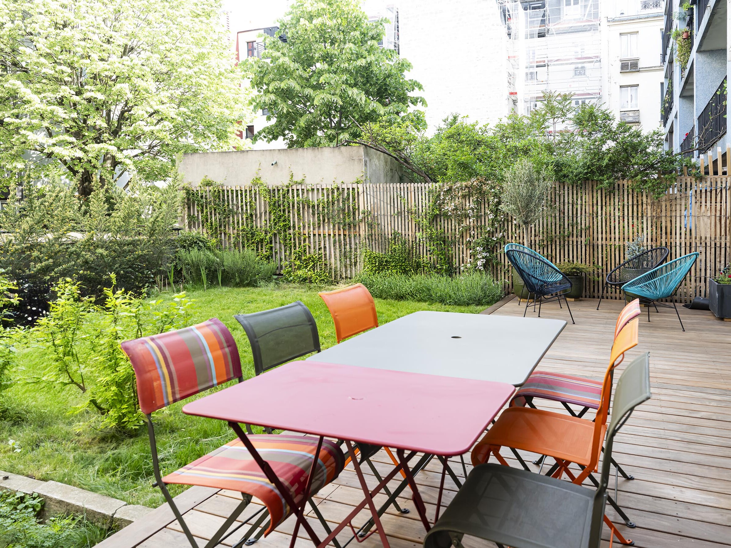 Un patio avec une table et des chaises colorées se trouve sur une terrasse en bois à côté d'un jardin vert avec des arbres et des plantes ; deux chaises longues se trouvent à l'arrière-plan.