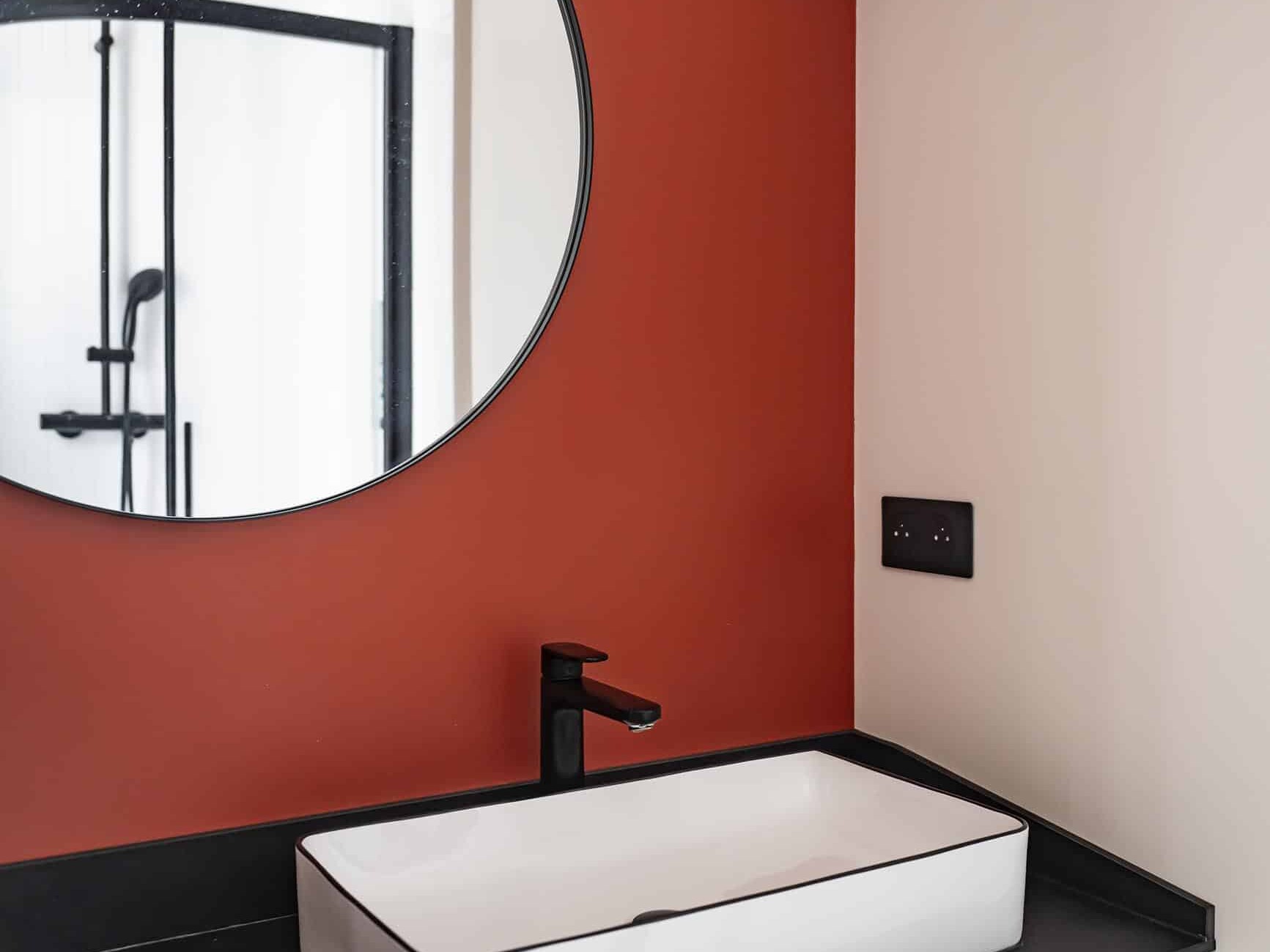 Salle de bains moderne avec un lavabo rectangulaire, un robinet noir, un miroir rond, un mur d'accent rouge et un plan de travail noir. Une douche avec une porte en verre est visible dans le reflet.
