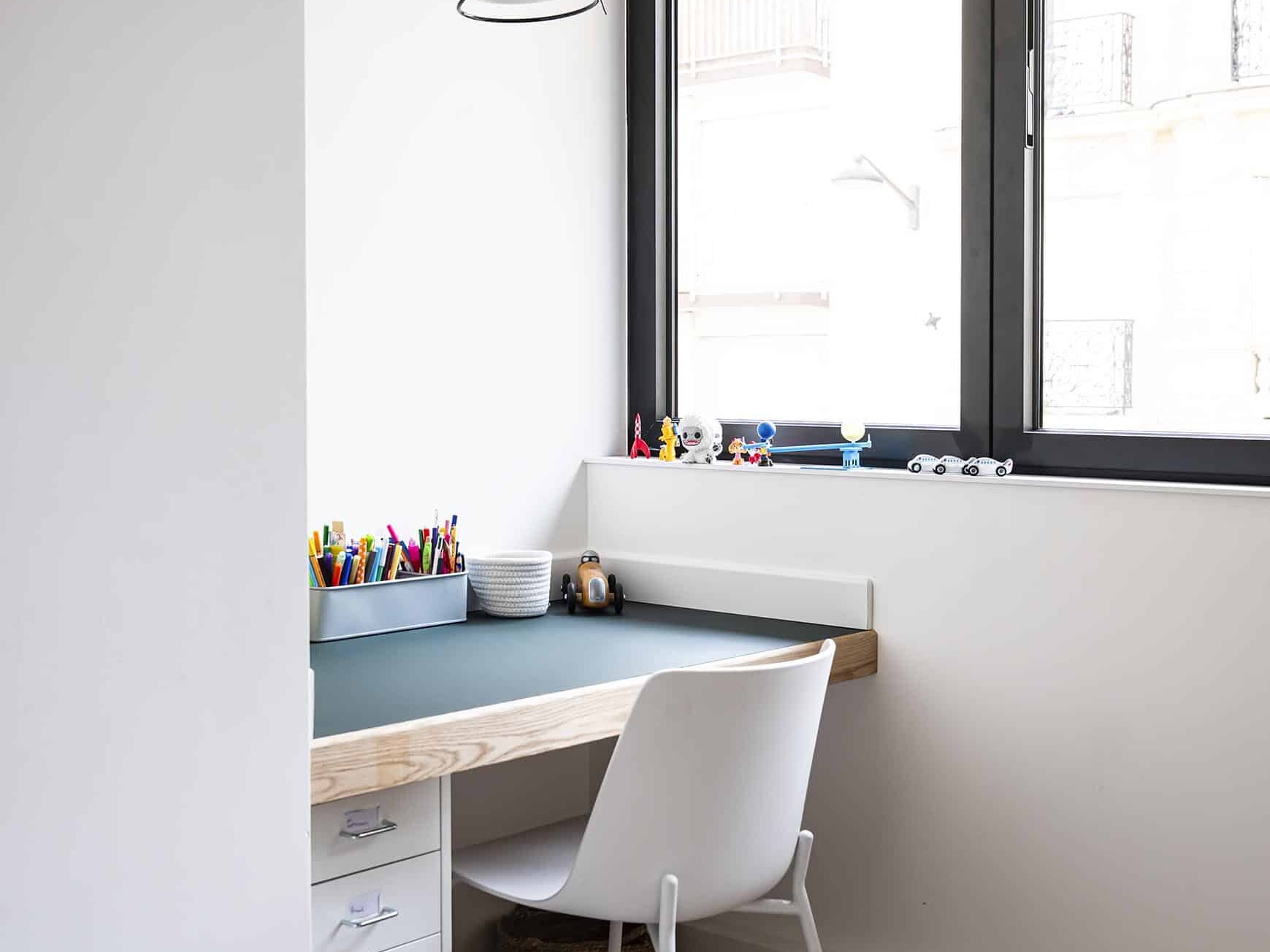 Un petit bureau organisé avec des fournitures de bureau et des jouets se trouve près d'une fenêtre aux cadres noirs ; une chaise blanche et une lampe orange sont placées à proximité.