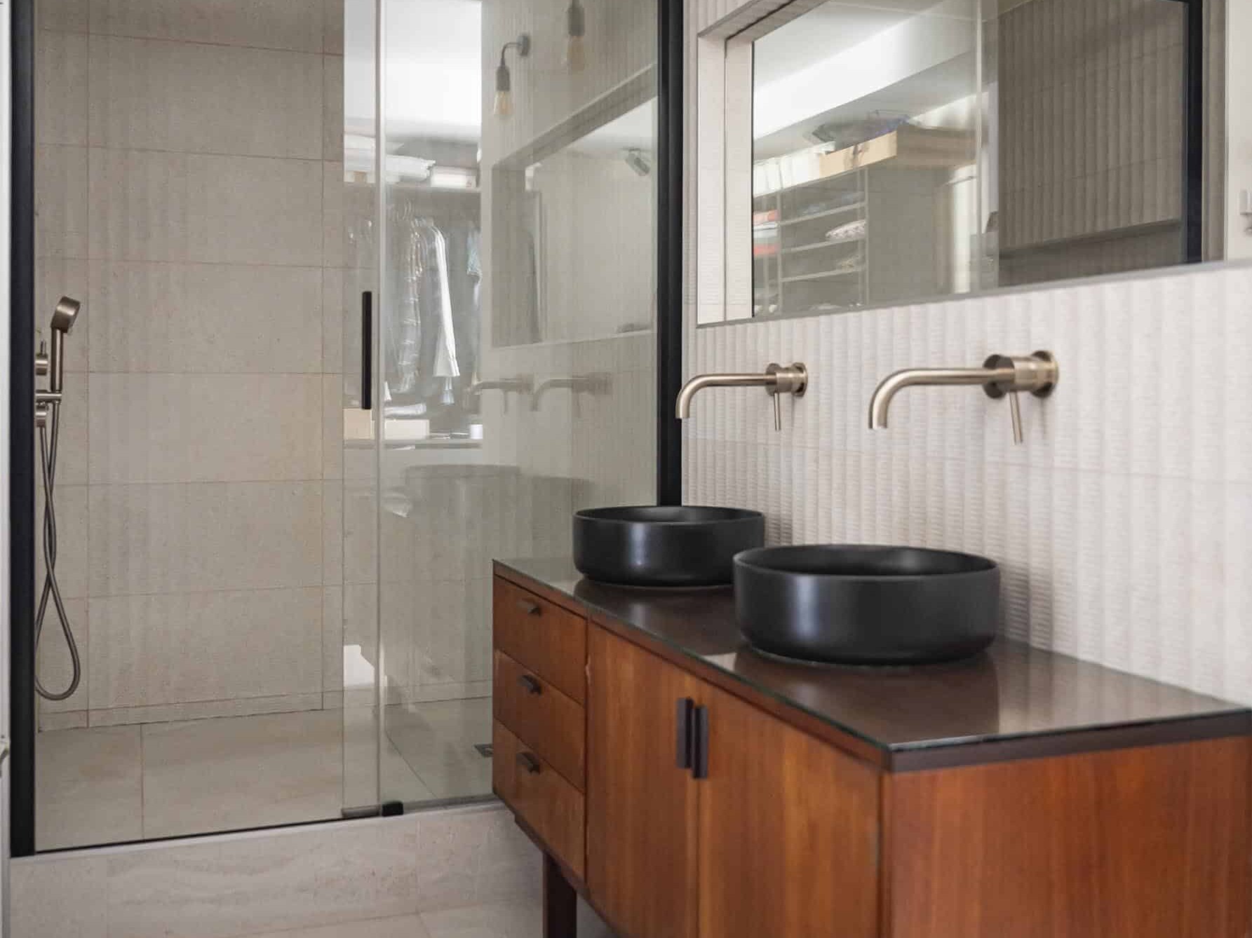 Salle de bains moderne avec douche vitrée, double lavabo avec vasques noires, meuble-lavabo en bois, grand miroir et appliques murales.