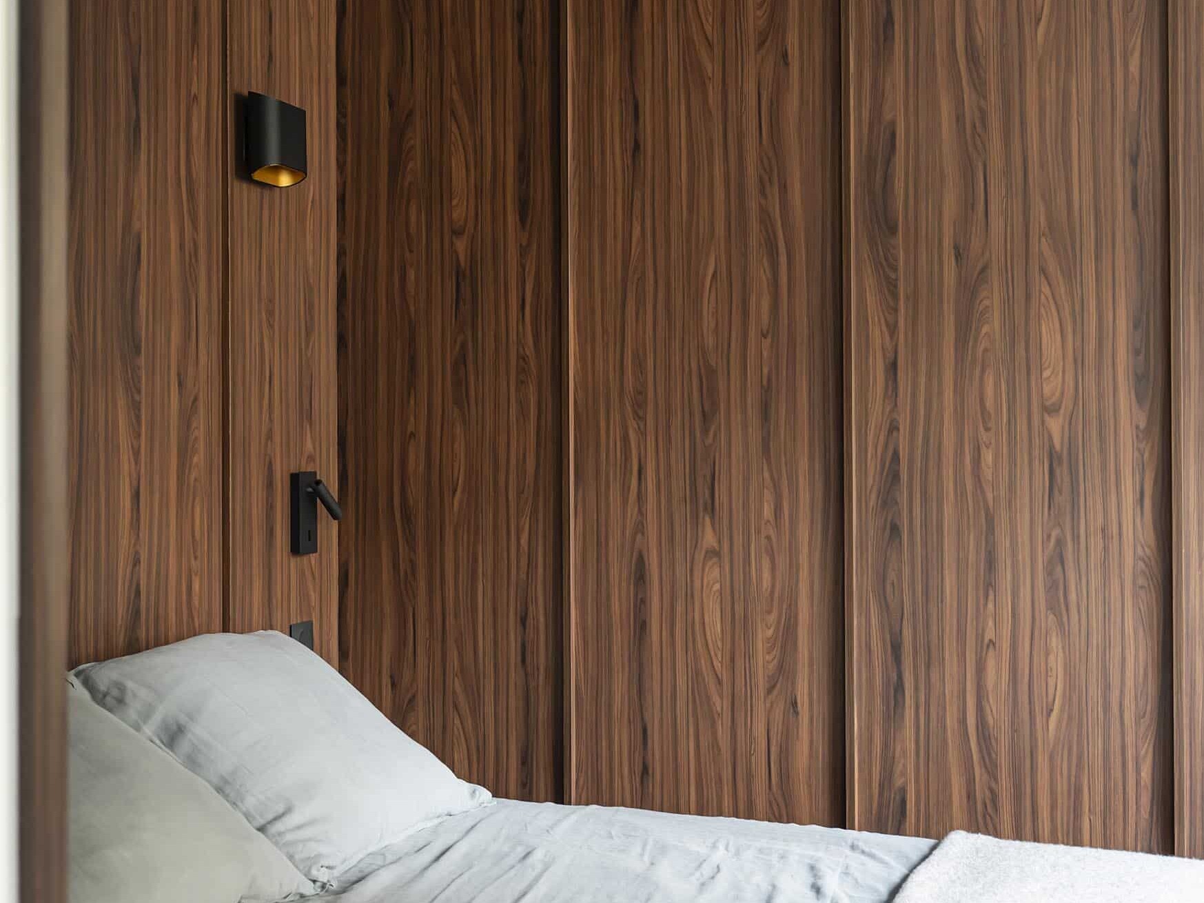 Une chambre à coucher minimaliste avec des murs et un plafond en bois, un lit simple avec une literie grise et une petite applique noire au-dessus du lit.