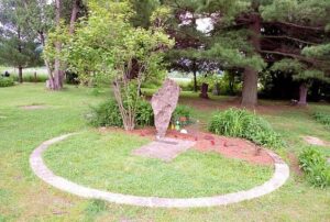 Un monument en pierre à peu près vertical se dresse à l'intérieur d'une petite bordure circulaire en pierre, entouré d'herbe, de buissons et d'arbres dans un cadre semblable à celui d'un parc - un écho de l'histoire de Paris, qui rappelle une photo ancienne.