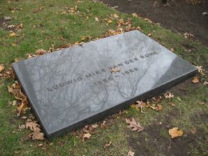 Une simple pierre tombale noire pour Ludwig Mies van der Rohe (1886-1969) repose à plat sur le sol, entourée d'herbe et de feuilles brunes éparses, comme une photo ancienne capturant un moment calme de l'histoire.