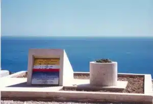 Une pierre tombale moderniste aux motifs géométriques colorés se dresse à côté d'une jardinière ronde surplombant une mer calme et bleue sous un ciel clair, évoquant l'élégance tranquille d'une photo ancienne de Paris et murmurant des échos de l'histoire.