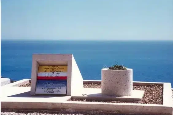 Une pierre tombale moderniste aux motifs géométriques colorés se dresse à côté d'une jardinière ronde surplombant une mer calme et bleue sous un ciel clair, évoquant l'élégance tranquille d'une photo ancienne de Paris et murmurant des échos de l'histoire.