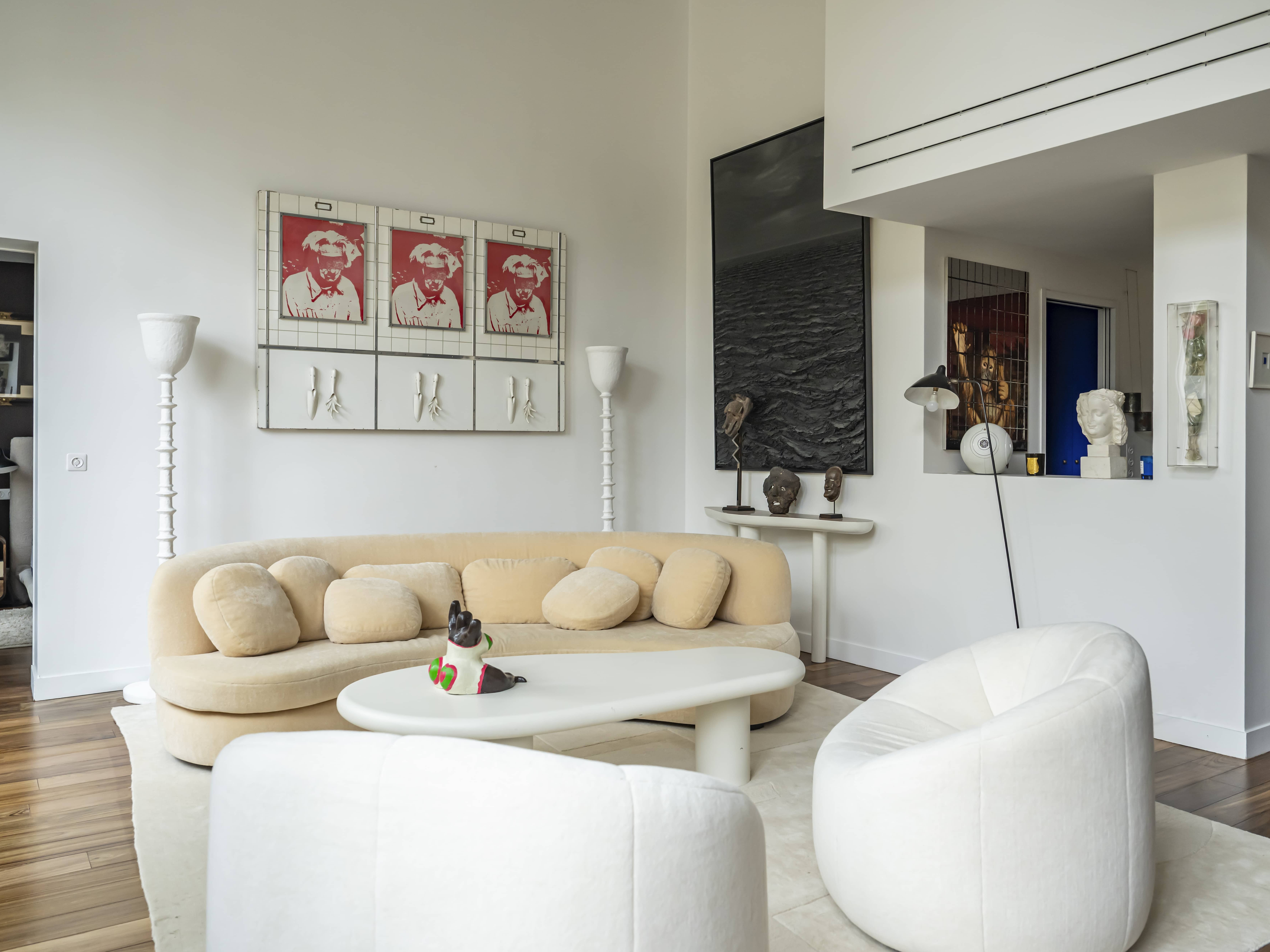 Un salon moderne dans un appartement familial haussmannien, avec des meubles beiges, deux chaises blanches, une table basse ronde et de l'art contemporain sur des murs blancs et un sol en bois.