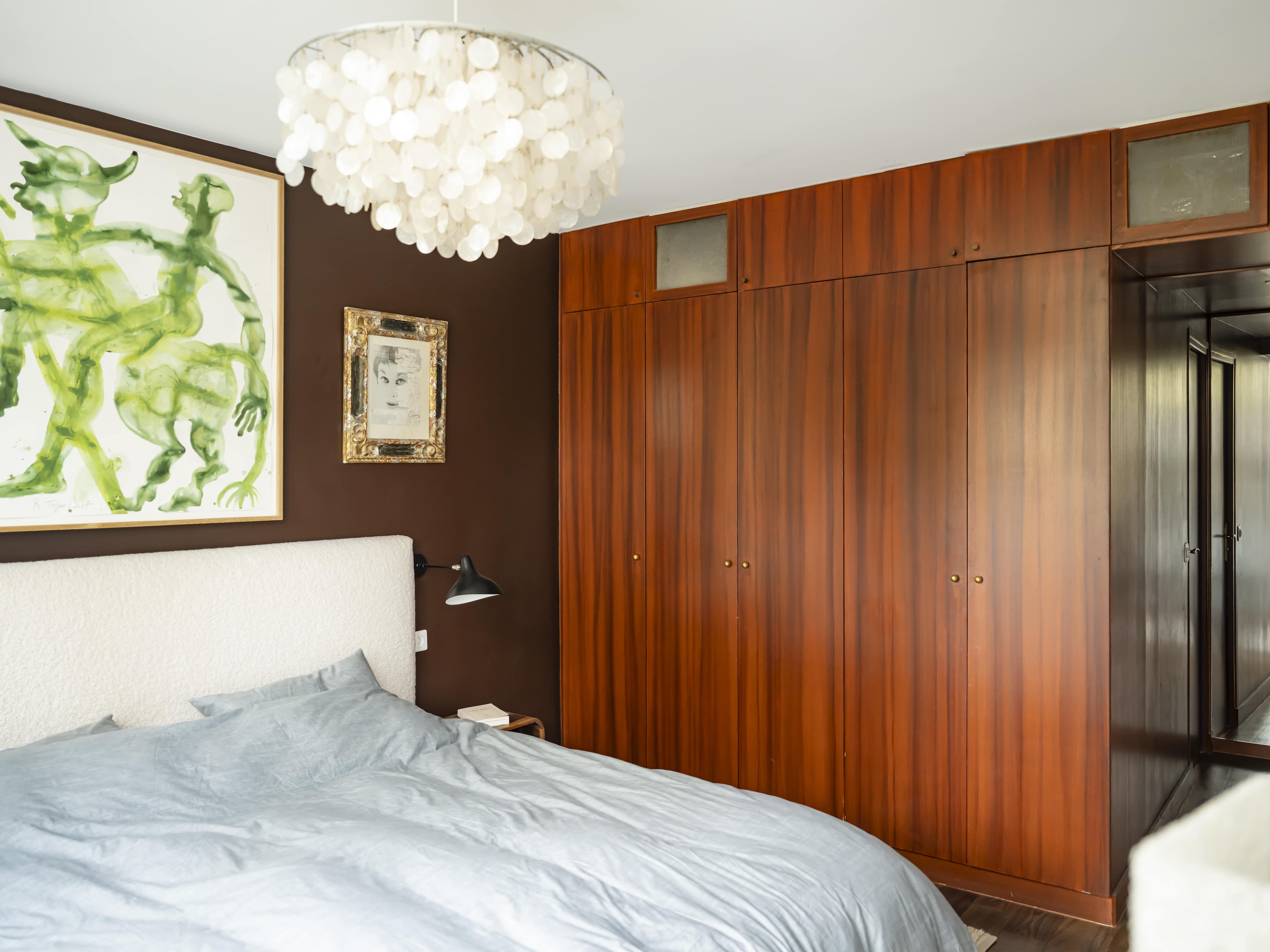Une chambre dans un appartement familial avec un lit gris clair, un mur marron foncé orné d'œuvres d'art abstraites vertes, des armoires encastrées en bois et un lustre en coquillage blanc.