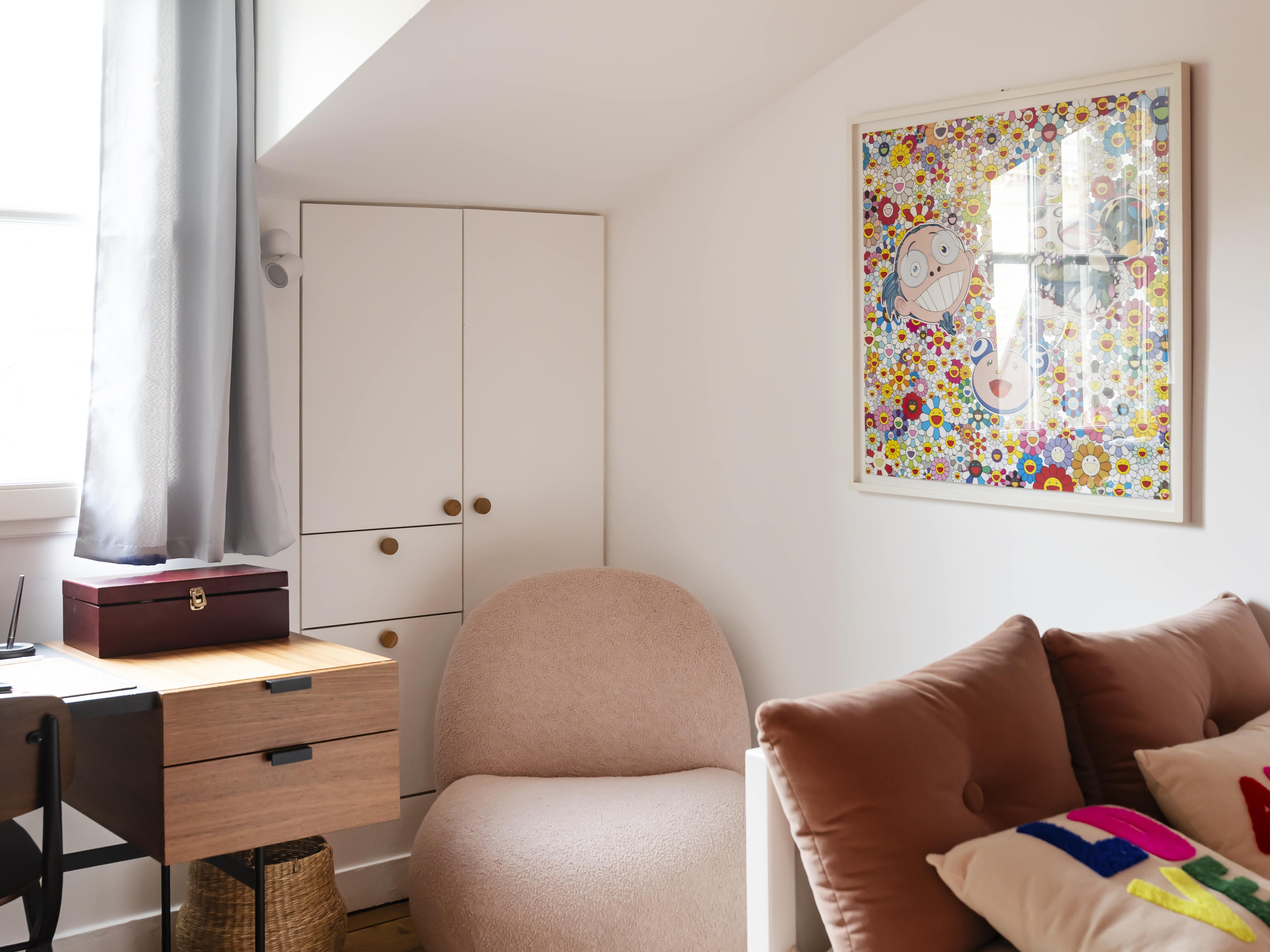 Un coin de chambre confortable avec une chaise longue rose, un lit avec des oreillers colorés, un bureau avec des tiroirs, une fenêtre avec des rideaux gris et une œuvre d'art encadrée colorée sur le mur.