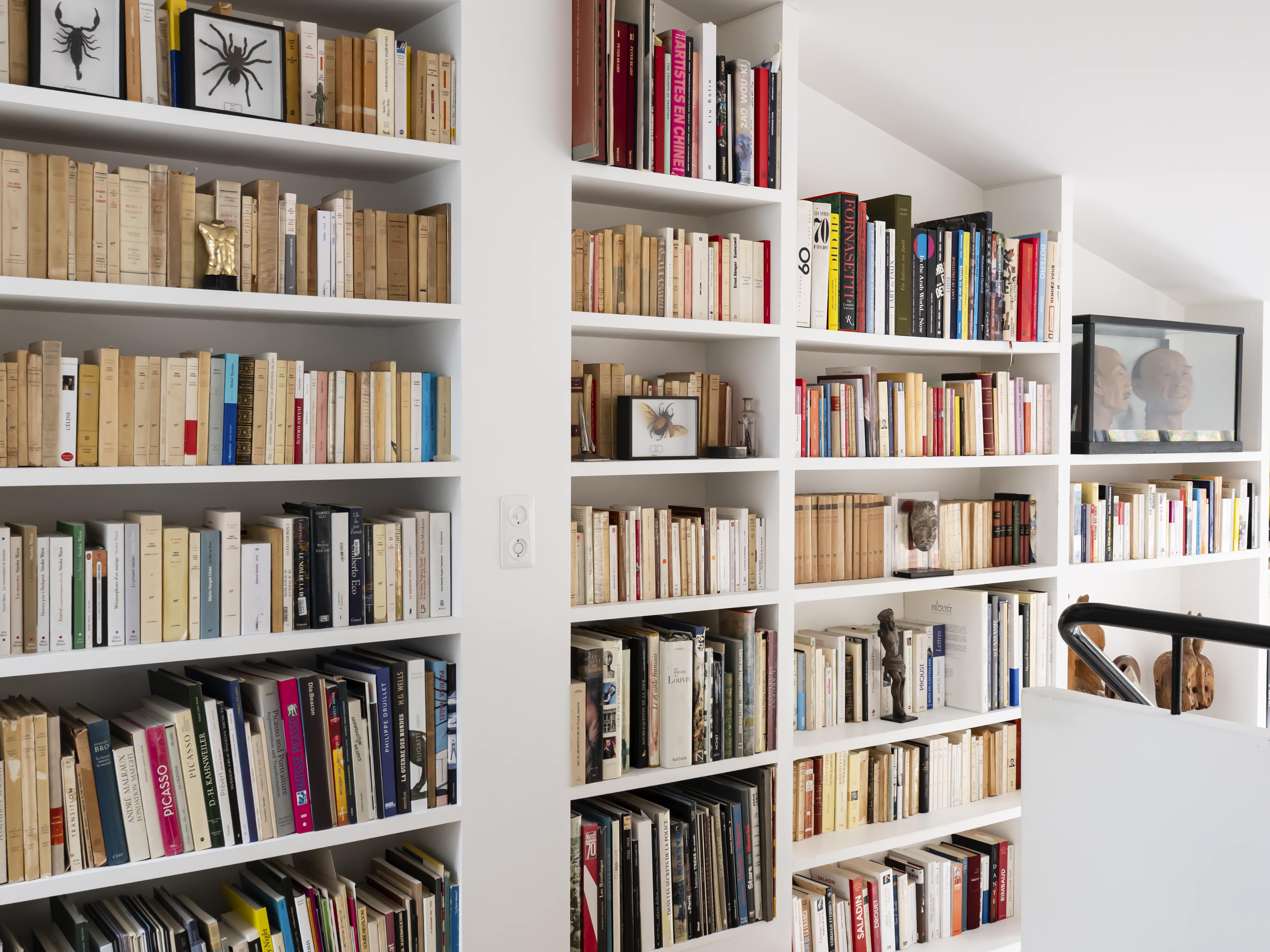 Des étagères remplies de livres tapissent les murs blancs de cet appartement familial moderne et lumineux, avec du parquet et un tapis blanc. Un tableau et deux dessins encadrés sont exposés sur les étagères.