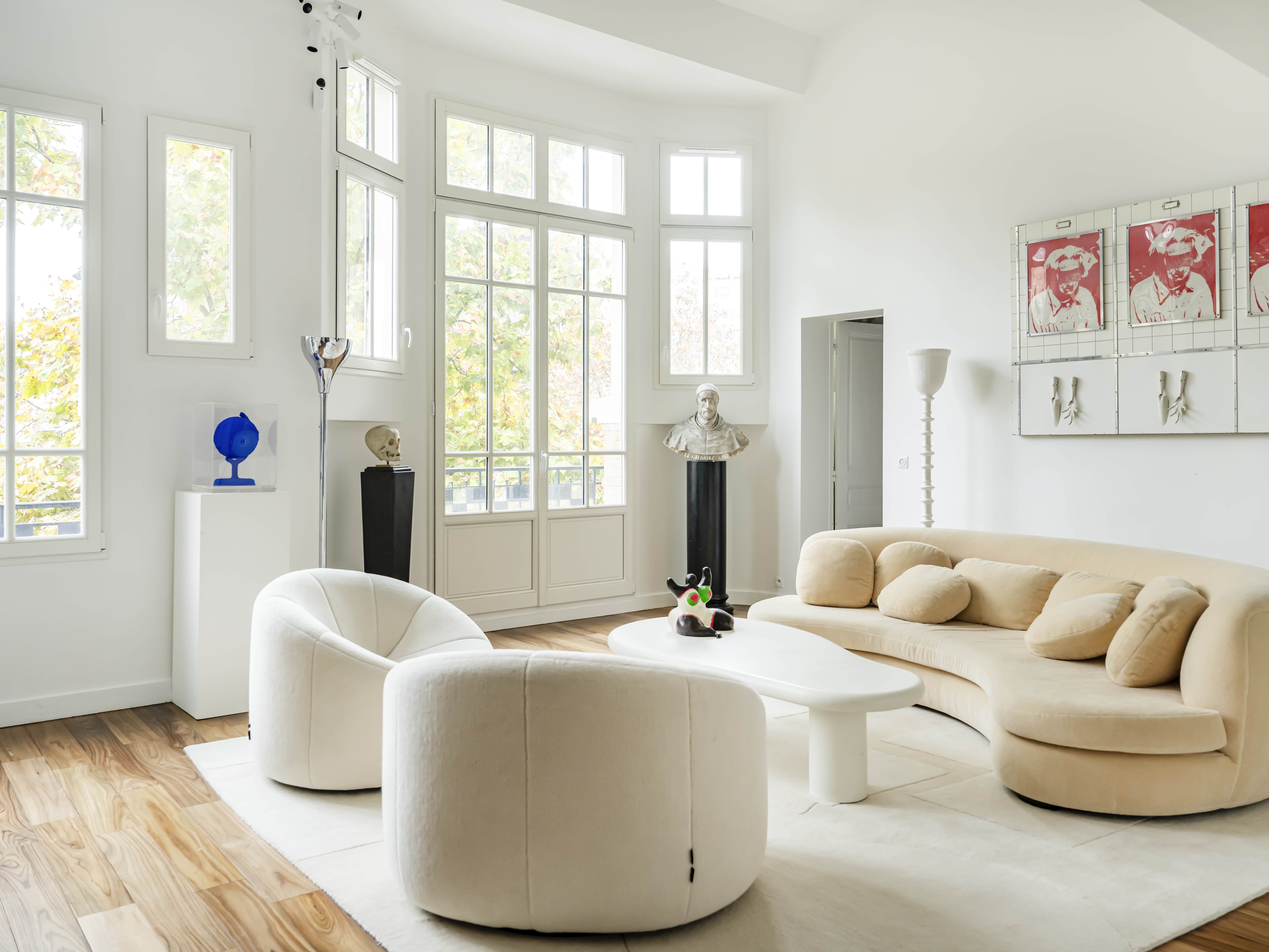 Salon haussmannien moderne avec des meubles incurvés de couleur crème, des murs blancs, de grandes fenêtres, des œuvres d'art et du parquet. Cet appartement familial est baigné de lumière naturelle et dispose d'une chambre en plus pour plus de confort.