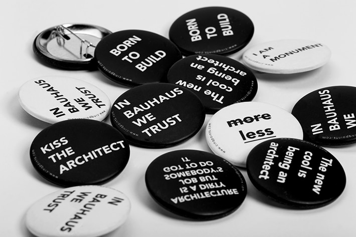 Une collection de boutons noirs et blancs avec diverses phrases liées à l'architecture telles que "IN BAUHAUS WE TRUST" et "BORN TO BUILD" dispersées sur une surface blanche.