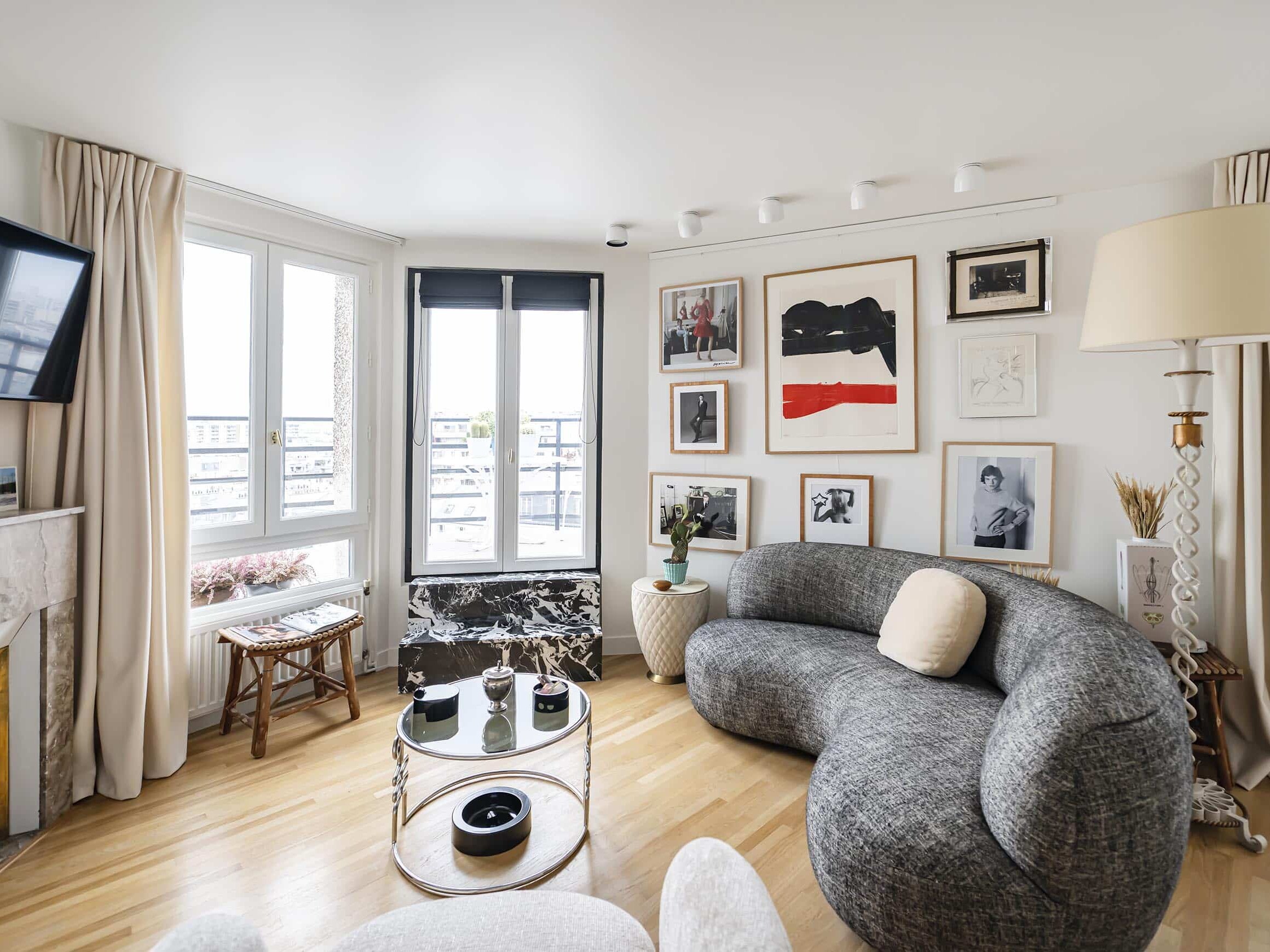 Salon haussmannien moderne avec un canapé gris incurvé, des tables basses rondes, des œuvres d'art sur des murs blancs, de grandes fenêtres avec des rideaux beiges et des planchers en bois - idéal pour un appartement familial élégant.