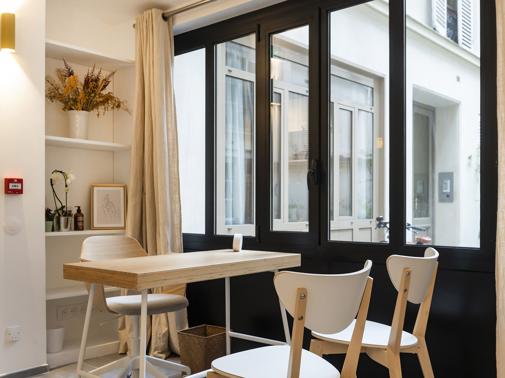 Un espace de travail moderne et minimaliste qui rappelle un cabinet médical élégant, composé d’une table en bois, de chaises blanches, d’étagères décorées et de grandes fenêtres ornées de rideaux beiges laissant entrer la lumière naturelle — idéal pour ceux qui cherchent l’inspiration au milieu de travaux de rénovation récents.
