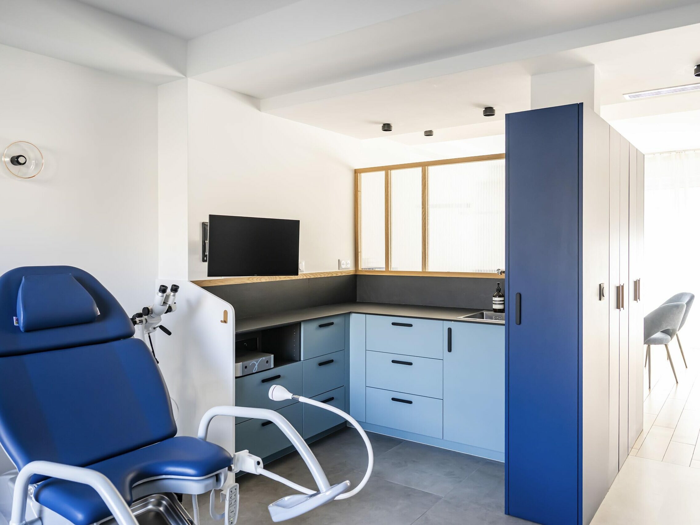 Salle d'examen médical moderne équipée d'un fauteuil d'examen bleu, d'armoires, d'un petit lavabo, d'un écran mural et bénéficiant d'un éclairage naturel abondant : un espace conçu avec soin par un architecte spécialisé dans la rénovation à Paris.