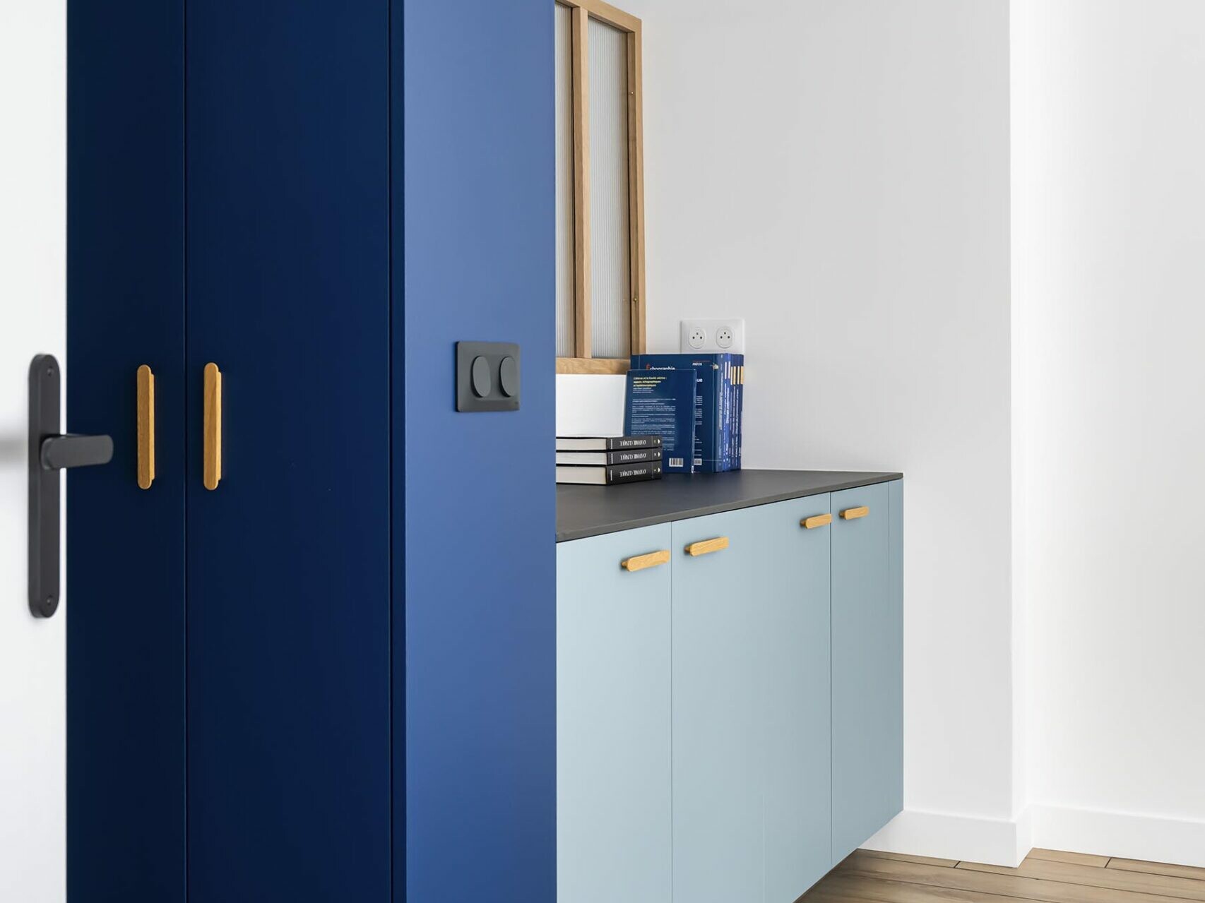Cuisine moderne avec une armoire haute bleue, des meubles bas bleu clair, des poignées en bois, un plan de travail noir et un parquet. On aperçoit les murs et le plafond blancs, rehaussés de luminaires noirs, qui créent une ambiance raffinée rappelant celle d’un appartement familial haussmannien.