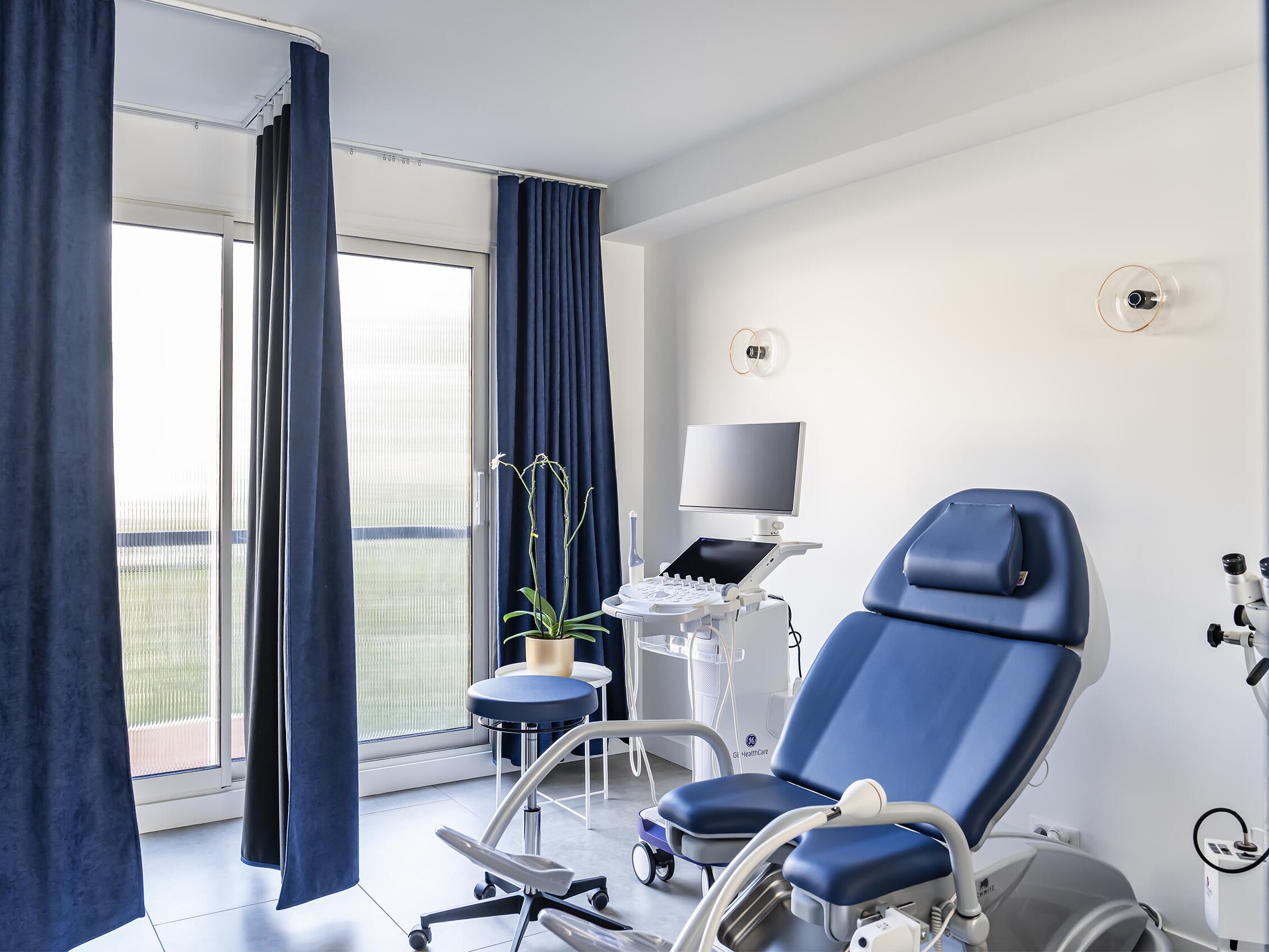 Salle d'examen médical équipée d'une chaise bleue, d'un écran d'ordinateur, d'un échographe, d'un tabouret, d'une plante, de murs blancs et de grandes fenêtres ornées de rideaux bleu foncé — qui évoque la lumière raffinée que l'on pourrait trouver dans un appartement familial haussmannien doté d'une chambre supplémentaire.