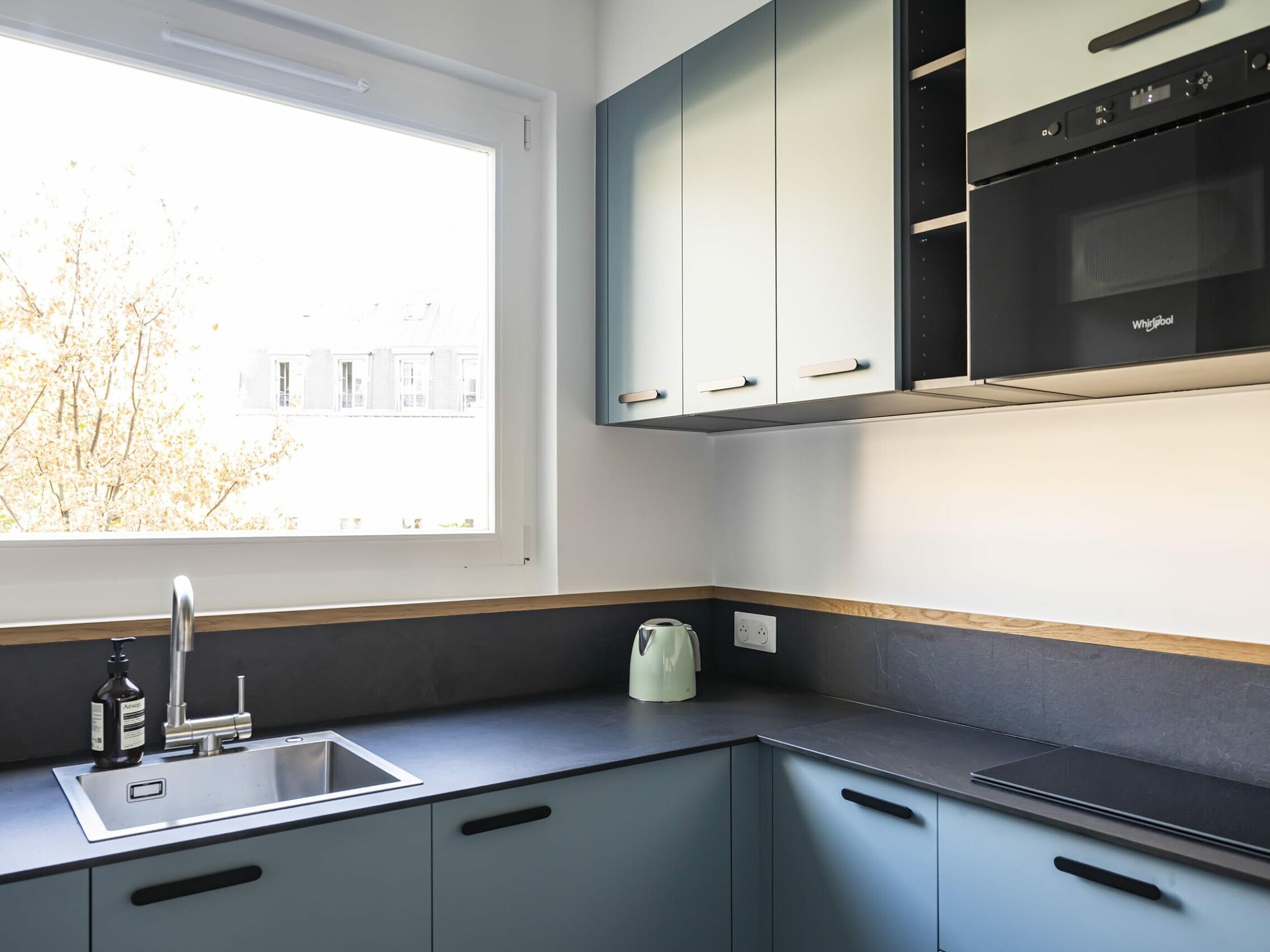 Cuisine moderne avec des placards bleus, un plan de travail noir, un évier, une bouilloire électrique verte et un four encastré sous les placards supérieurs, près d'une grande fenêtre — idéale pour un appartement familial ou pour quiconque recherche une chambre supplémentaire dans un espace au design raffiné.