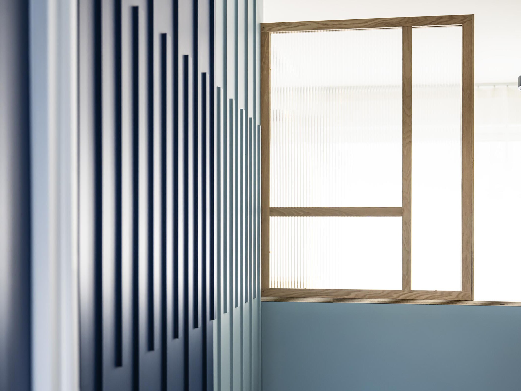 Un intérieur moderne avec un mur bleu orné de panneaux verticaux et une cloison vitrée à cadre en bois près d'une fenêtre, créant l'ambiance idéale pour une chambre supplémentaire dans un élégant appartement familial haussmannien.