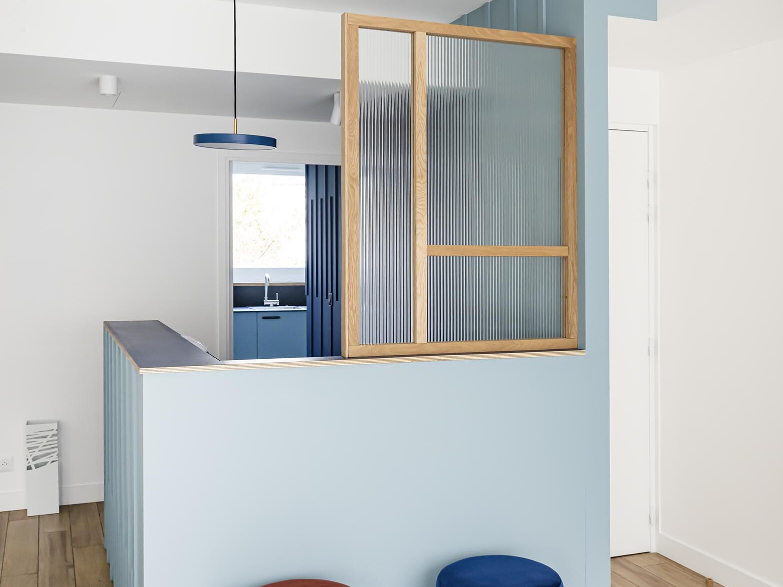 Intérieur haussmannien moderne avec une cloison bleue, des touches de bois, du verre nervuré et deux tabourets ronds en velours — l’un rouge, l’autre bleu — posés sur un parquet clair. La décoration minimaliste, associée à un éclairage doux et naturel, crée l’ambiance idéale pour un appartement familial à la recherche d’une chambre supplémentaire.