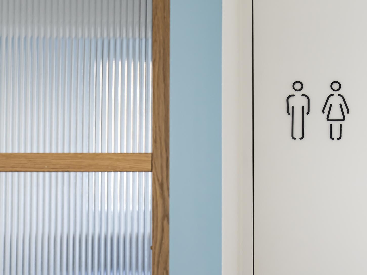 Une porte blanche ornée des symboles noirs des toilettes pour hommes et pour femmes, adossée à un mur bleu et à une fenêtre en verre dépoli encadrée de bois, rappelant les touches classiques que l'on retrouve dans un appartement familial haussmannien.