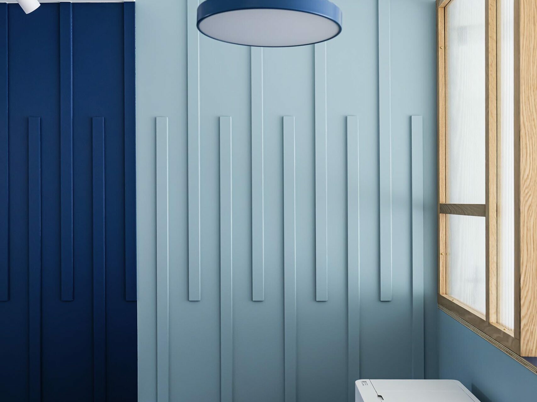 Intérieur minimaliste et moderne, avec des murs lambrissés de bleu, une suspension circulaire bleue, un plan de travail bleu et une fenêtre à encadrement en bois sur la droite — idéal pour un appartement familial haussmannien à la recherche d'une chambre supplémentaire ou d'une touche de modernité tout en conservant l'élégance parisienne classique.