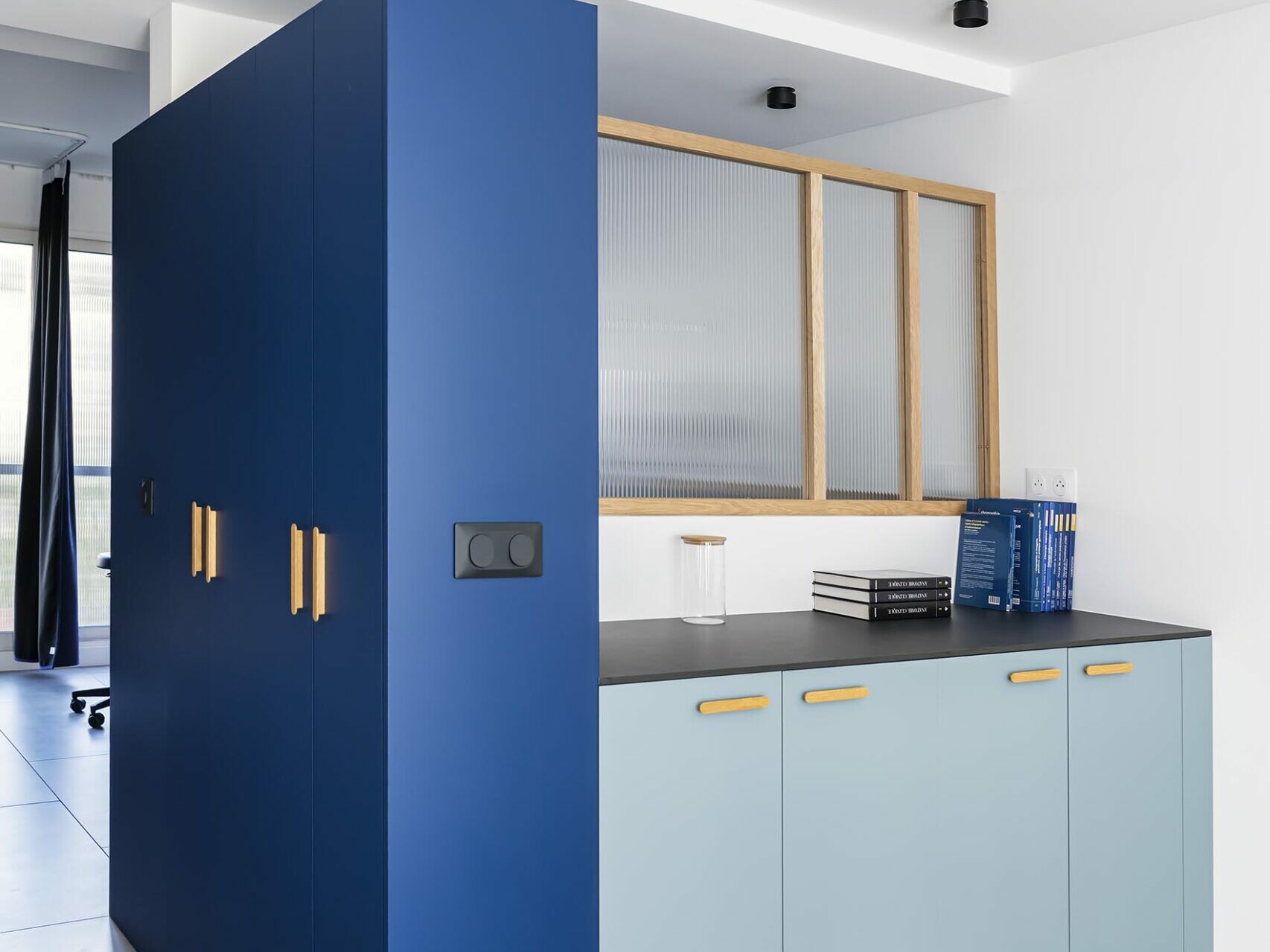 Un coin cuisine moderne avec des placards bleus et bleu clair, des poignées en bois, un plan de travail noir, un parquet et une cloison en verre dépoli — idéal pour un appartement familial situé dans un immeuble haussmannien chic.