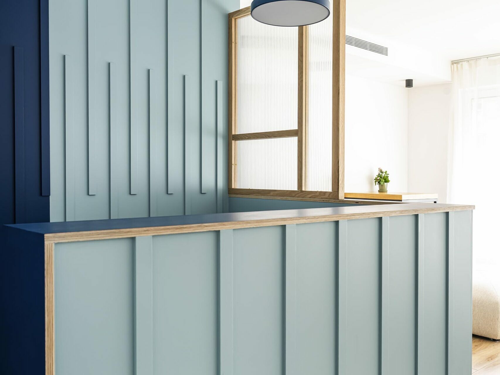 Une cloison moderne, composée de panneaux bleus verticaux et de moulures en bois, sépare deux espaces dans une pièce haussmannienne lumineuse, dotée d'un parquet et d'une suspension ronde au plafond, créant ainsi une circulation harmonieuse, idéale pour un appartement familial.