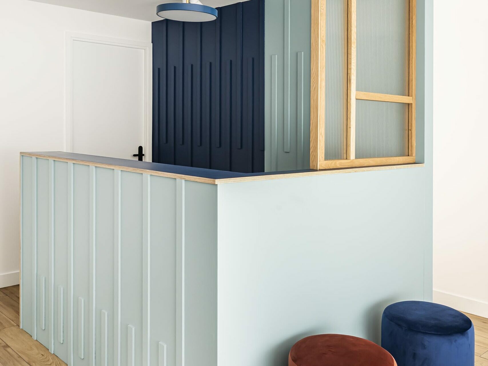 Un espace d'accueil moderne avec un comptoir lambrissé bleu pastel, un mur d'accent bleu marine, des moulures en bois, une suspension et deux tabourets ronds en velours — l'un rouge, l'autre bleu — posés sur un parquet clair ; une touche parfaite pour un appartement familial à la recherche d'une chambre supplémentaire ou d'un espace de vie polyvalent.