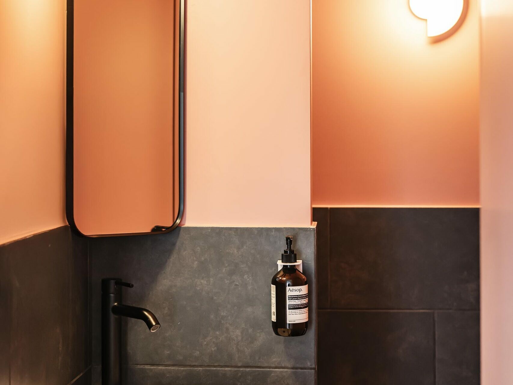 Salle de bains moderne avec des murs couleur pêche, un lavabo rond blanc, un robinet mural, un miroir rectangulaire, un distributeur de savon, du carrelage noir, des toilettes et deux appliques murales en forme de globe — idéale pour un appartement familial haussmannien recherchant le confort avec une chambre supplémentaire.